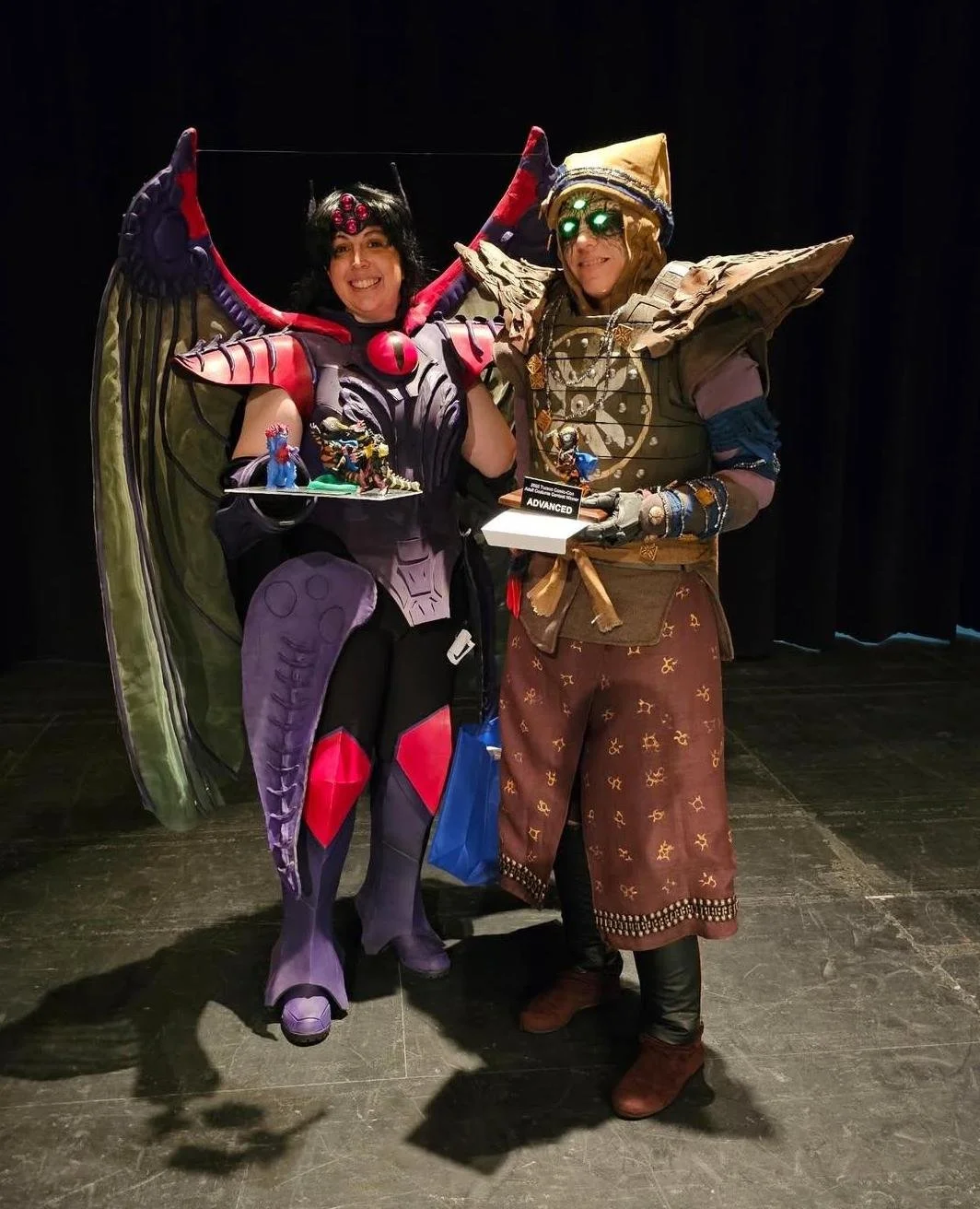 Eris Morn Cosplayer with 3 glowing green eyes and brown armor and dress, standing next to a Cosplayer in red and purple armor with dragon wings. They each hold a small trophy and they are smiling proudly. The background is dark