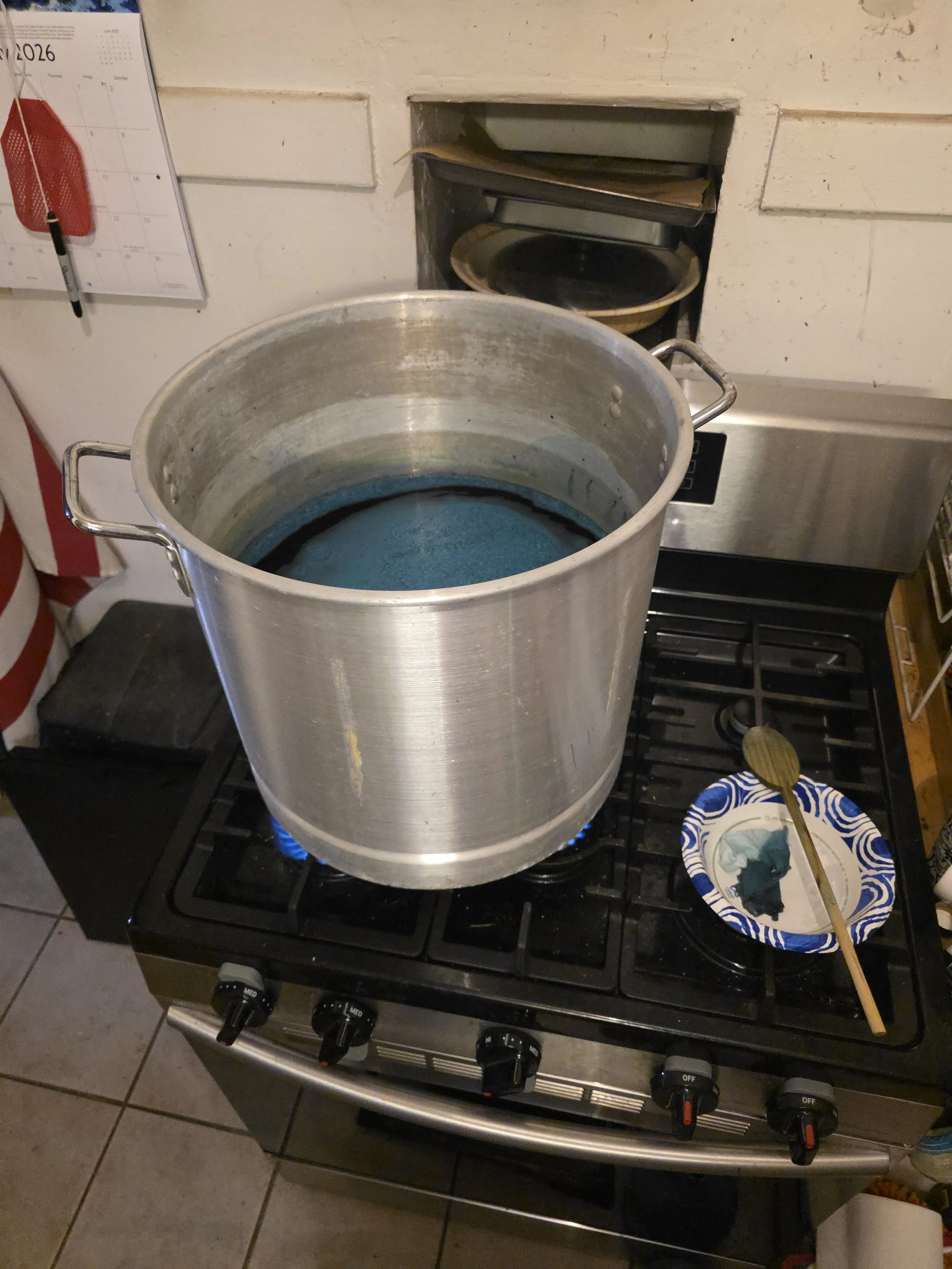 53 quart tamale pot sitting on 3 of the 5 burners on a gas stove. It is more than half full of dark green dye, and has fabric boiling in it and a wooden spoon resting in a paper bowl nearby