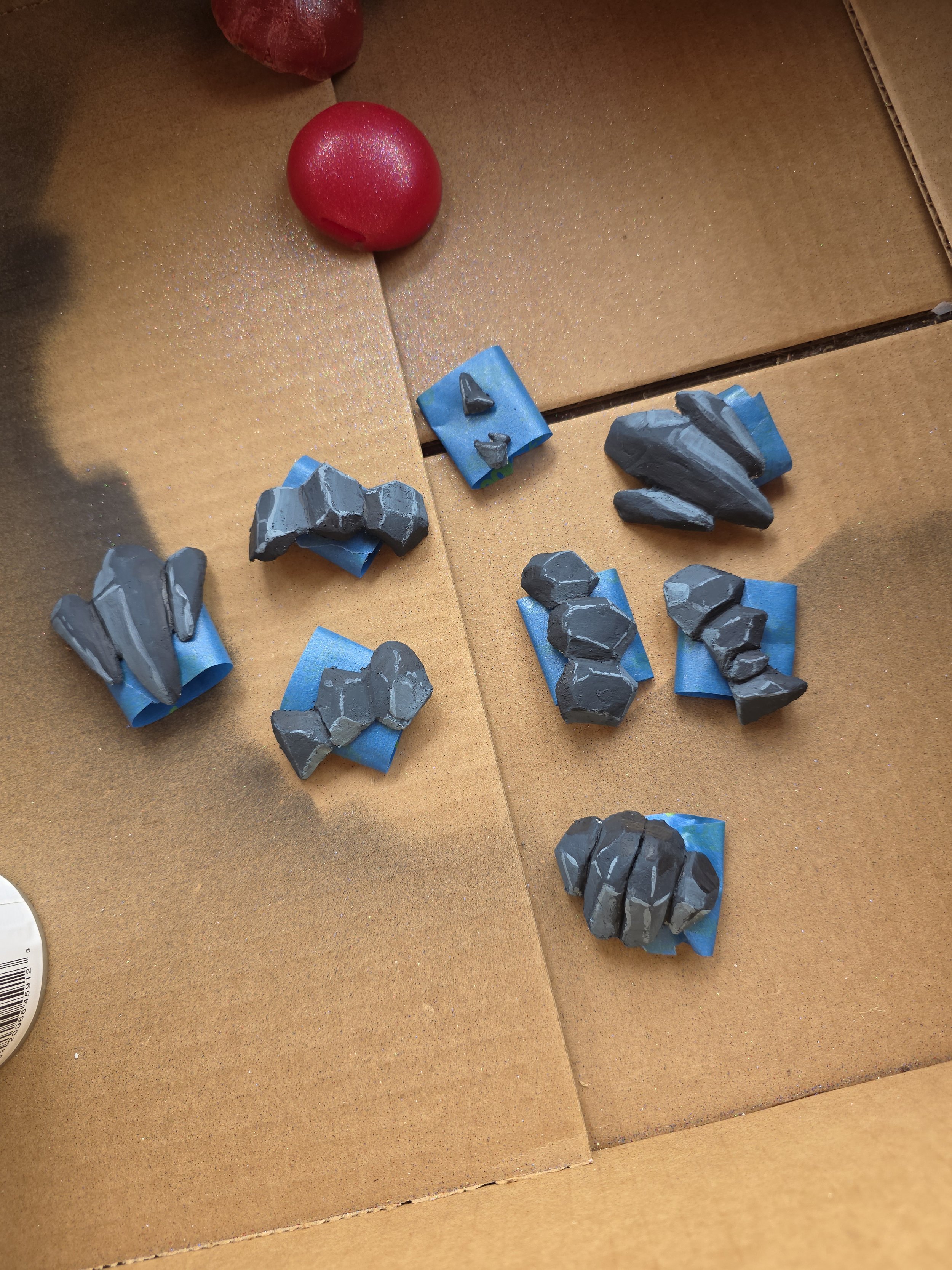 worbla rocks arranged in a cardboard box to catch spray paint overspray. Rocks are painted dark bluish gray with some lighter shading. They are taped down with blue tape rolled on the backs of them, and have a matte clear coat on them