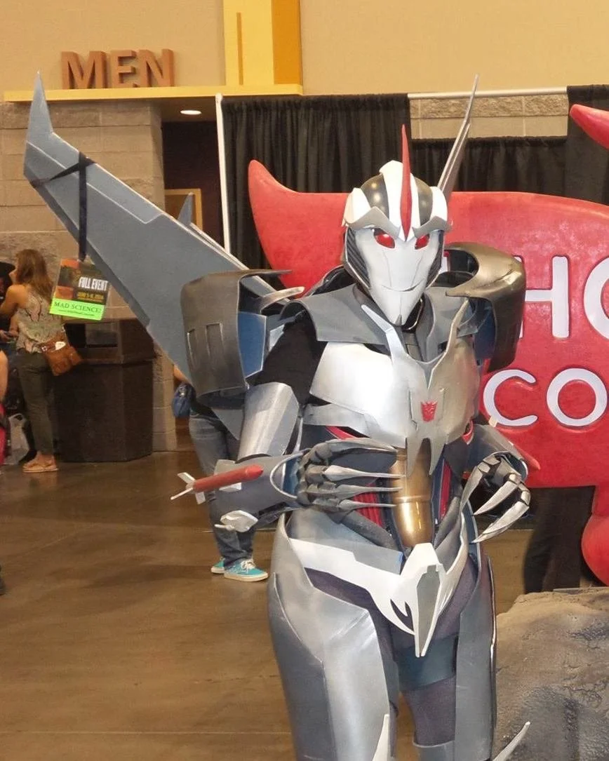 Cosplay of Transformers Prime Starscream, a slender robot with a pointed face and long jet wings. He has red details and eyes, with gray and silver armor. Both forearms have missiles attached. Picture is in a convention center