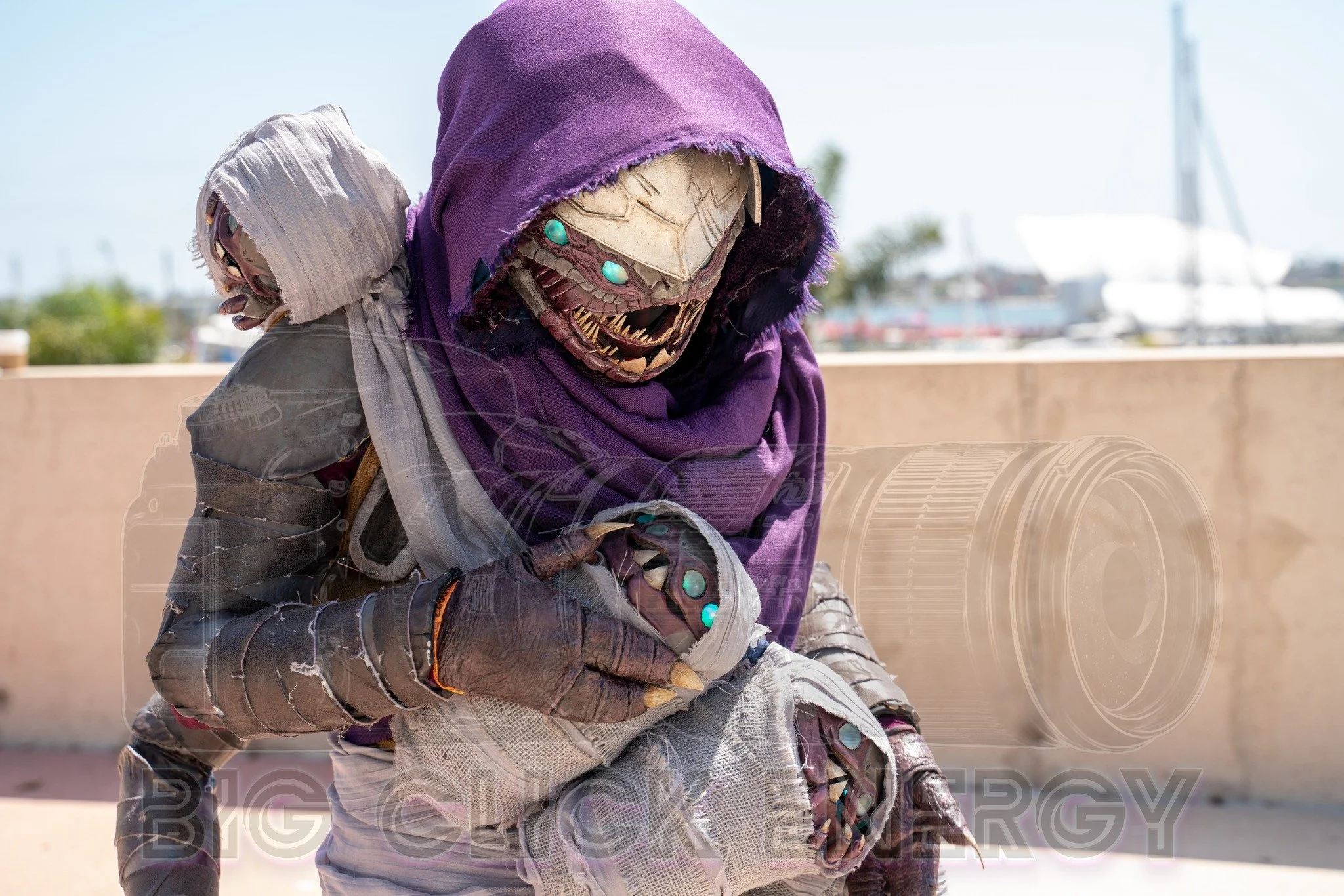 Cosplay of a Buglike alien in a purple hood with 4 glowing blue eyes, 2 claws on each hand, and 2 rows of needle-like teeth. It has 4 swaddled babies of the same species in carried in a baby sling