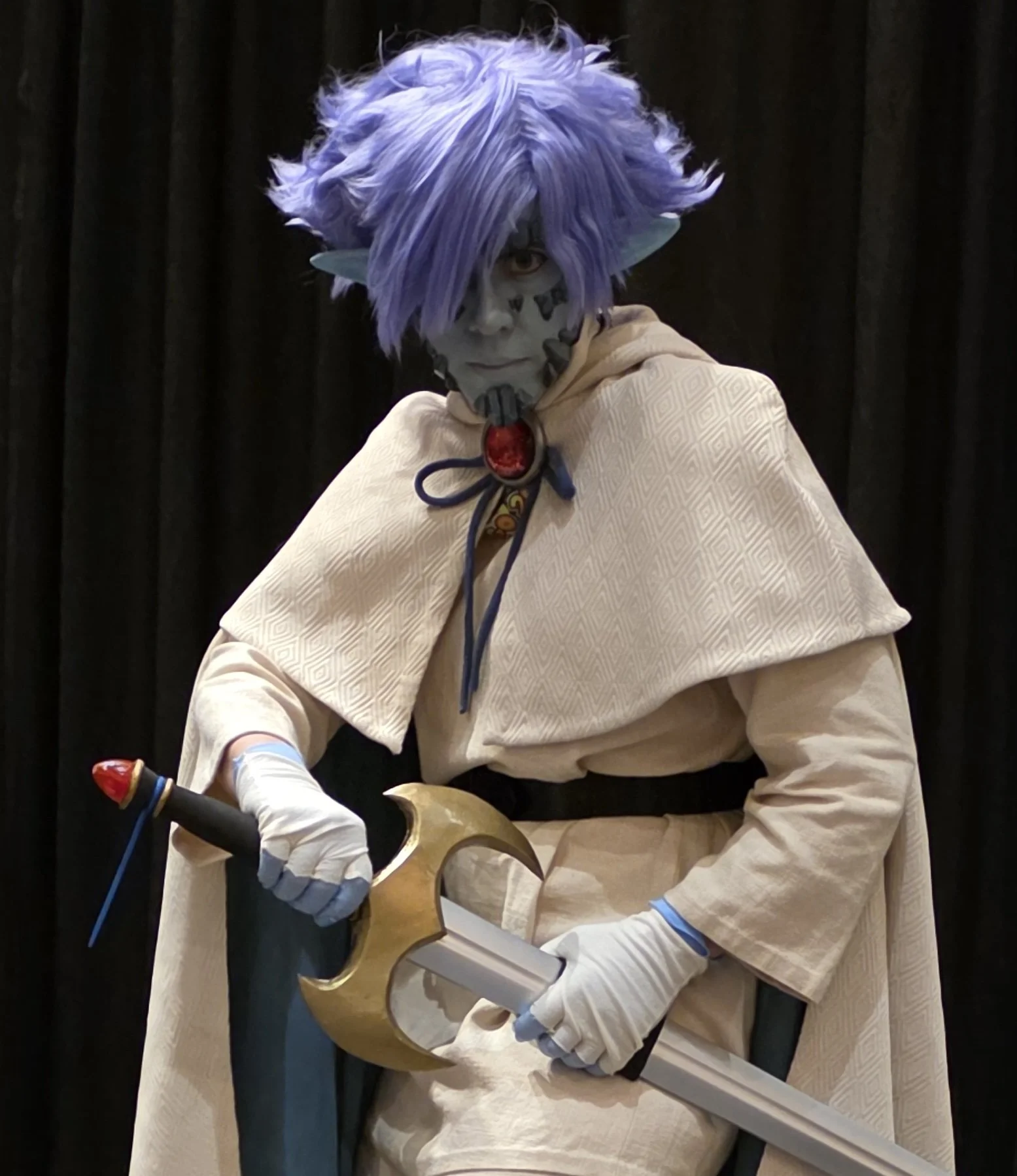 Cosplayer dressed as Zelgadis, an anime character in off-white pants and tunic, off-white cape with dark green lining, blue skin with scale-like rocks in his face, and lavender hair. He is about to draw a longsword with a large golden hilt