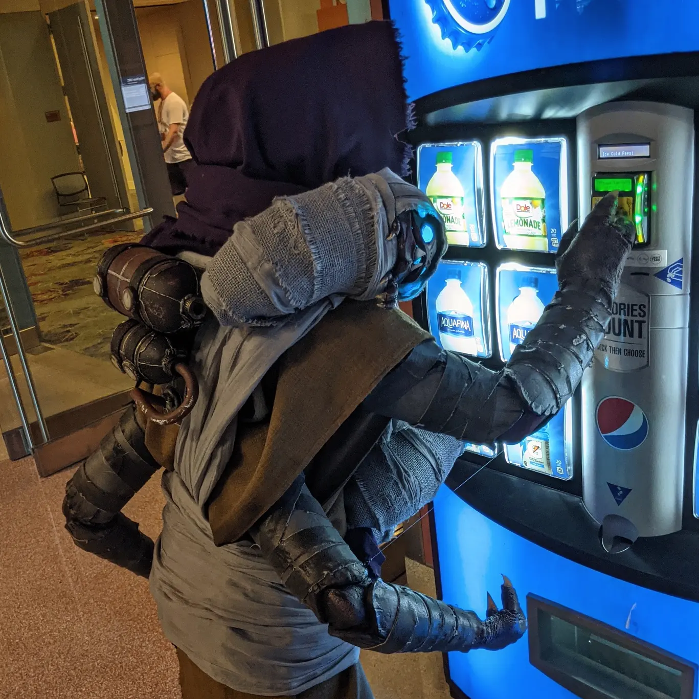 Cosplay of a Buglike alien in a purple hood with 4 glowing blue eyes, 2 claws on each hand, and 2 rows of needle-like teeth. It is trying to buy something from a Pepsi vending machine. It has 4 swaddled babies of the same species in carried in a baby