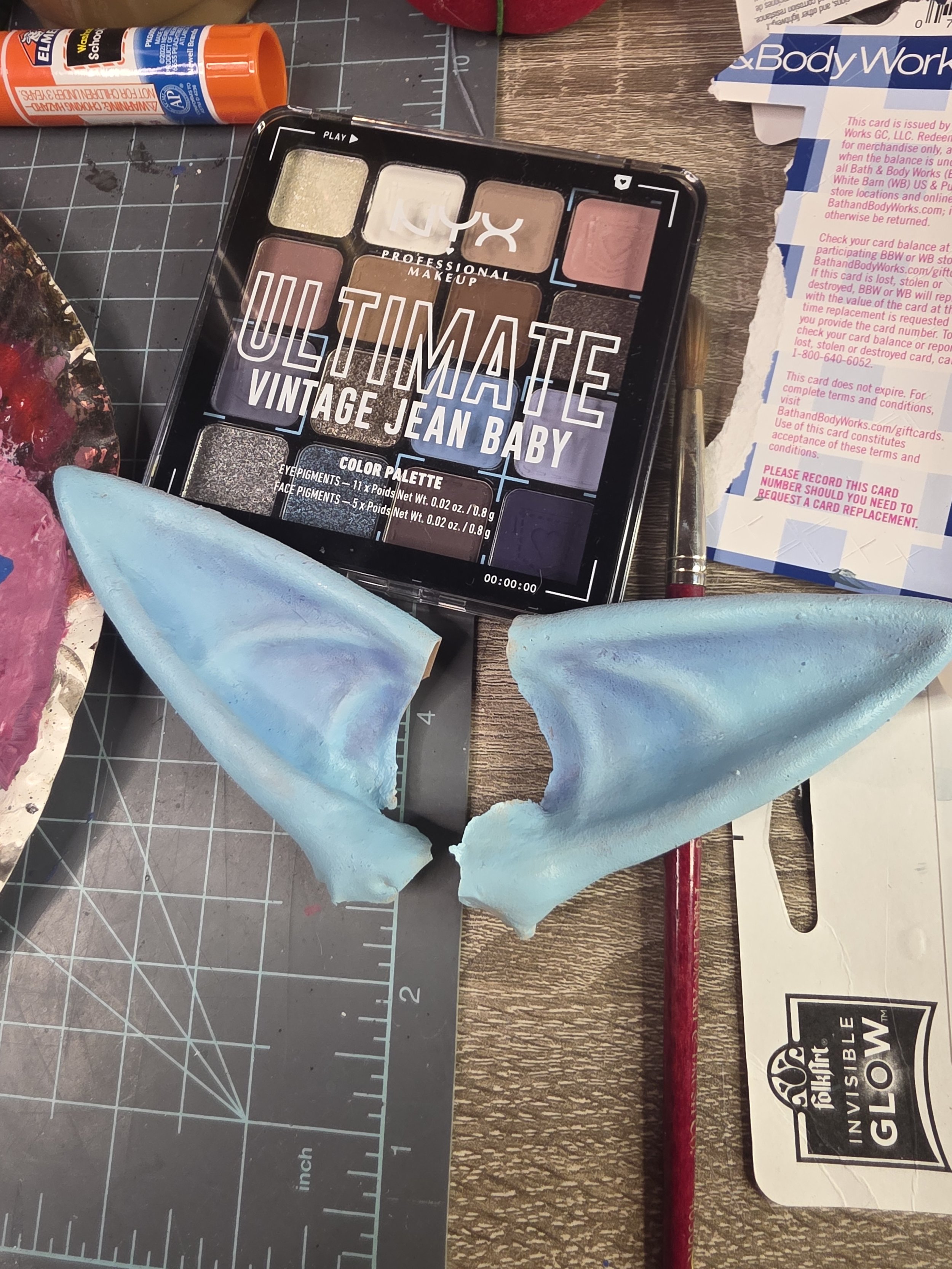 latex pointy ears, painted pale blue, sitting beside an eyeshadow pallet of browns, coppers, and blues. the anatomy of the ears is shaded with dark blue eyeshadow