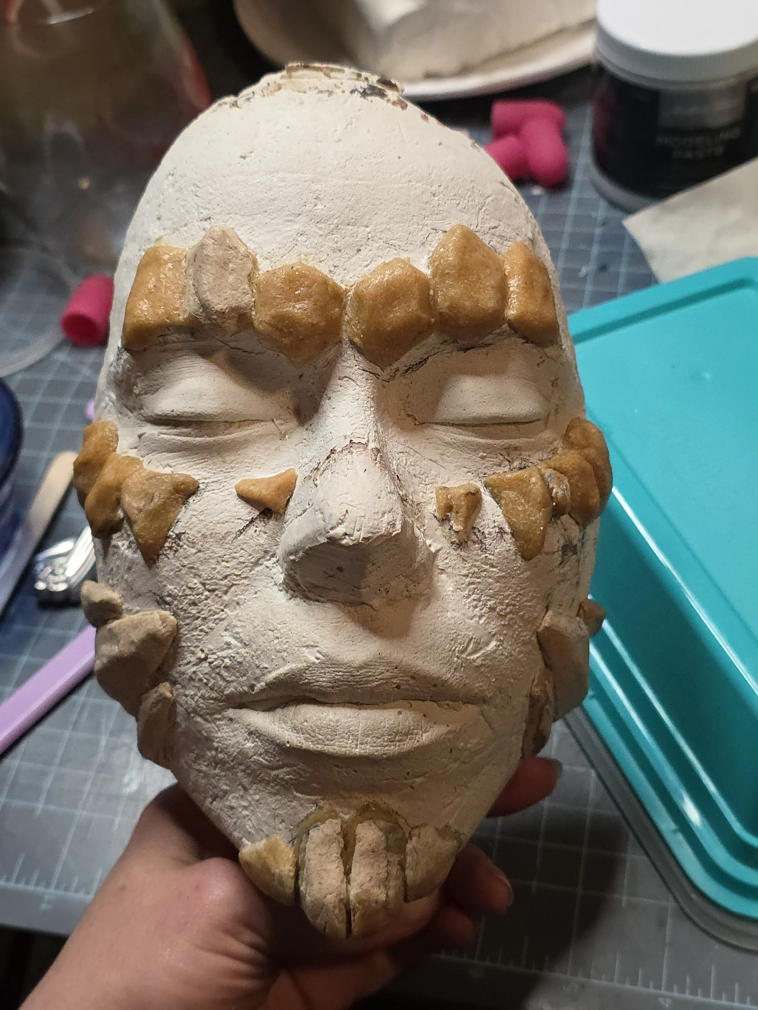 plaster cast of a face with pieces of tan colored Worbla stuck on to form rock-like scales. The eyebrows, cheekbones, and jawline have mostly flat scales with some built-up thickness, and the chin has a goatee like shape made up worbla rocks