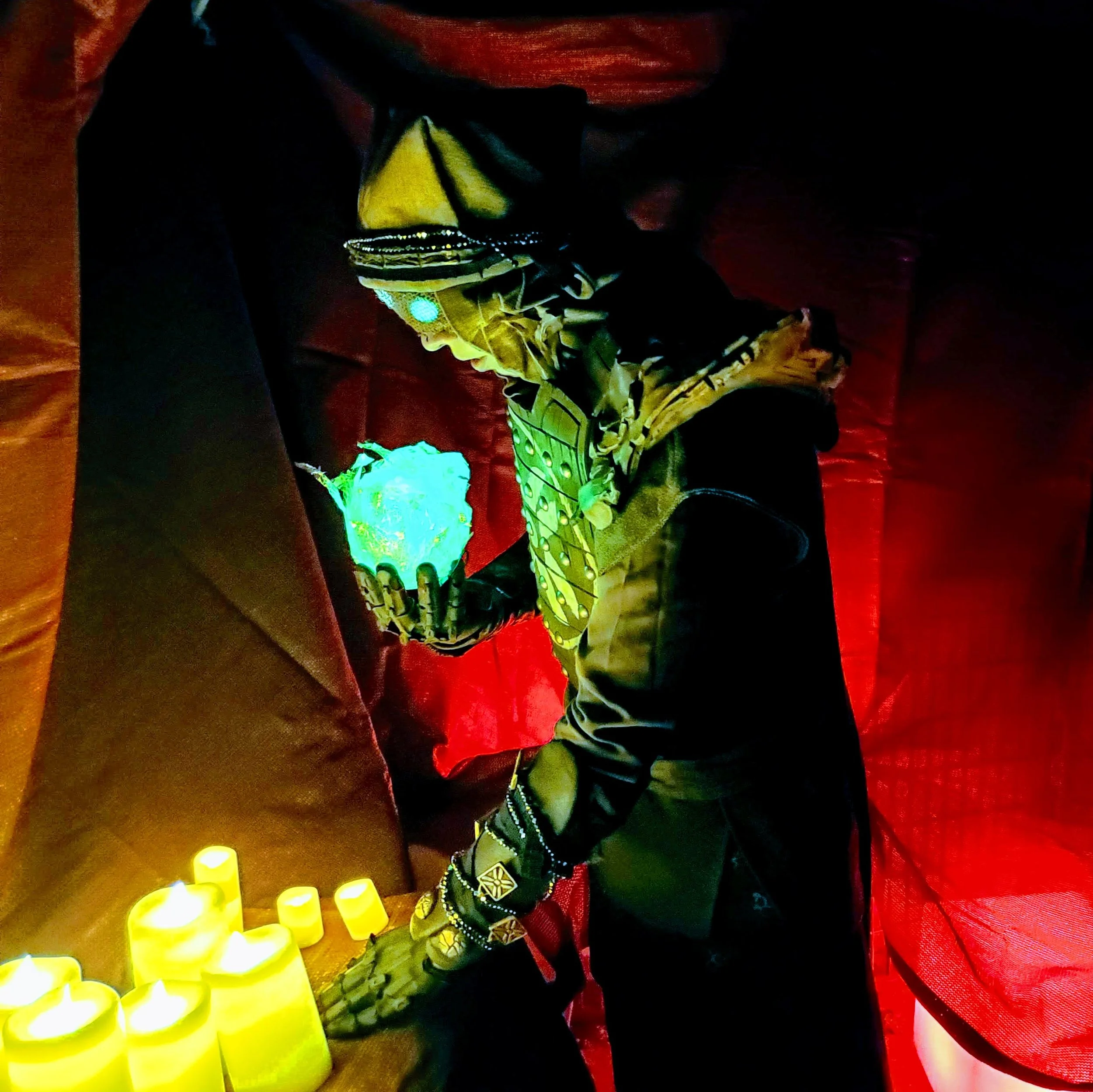 Eris Morn Cosplayer with 3 glowing green eyes holding a glowing green orb over an array of candles. She wears weathered brown armor and strings of beads and charms. The background is dimly lit and red