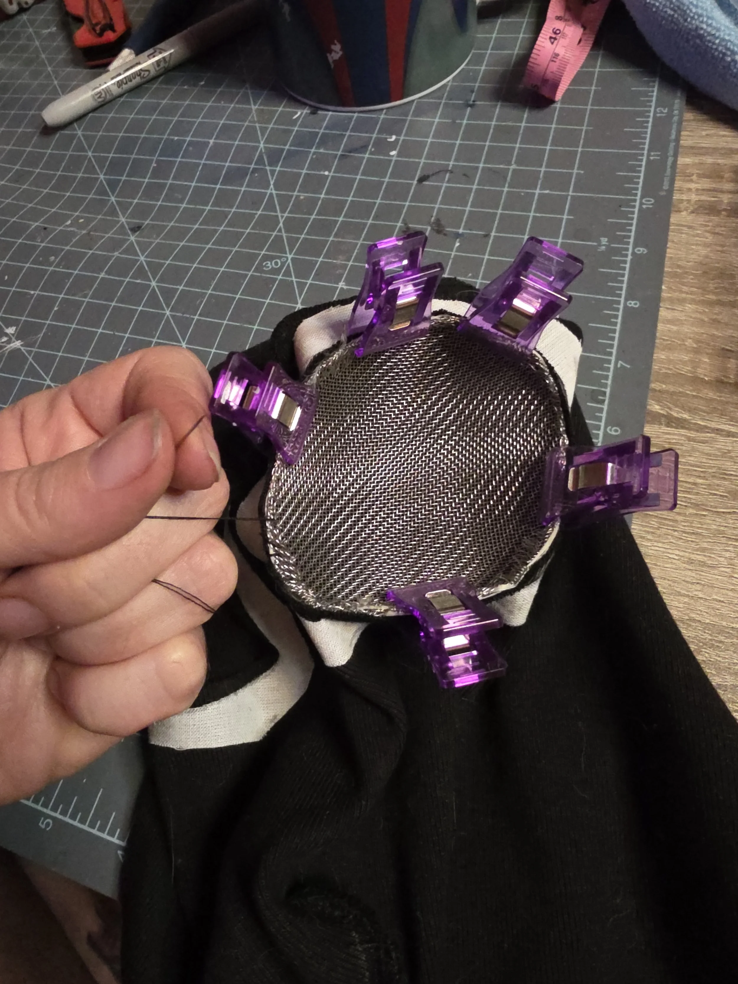 Metal mesh half egg shaped eye cover clipped into the eye hole of a black ninja mask with purple plastic clips. A thread is attached to the side of the hole and fingers are holding it to stitch the metal screen to the fabric