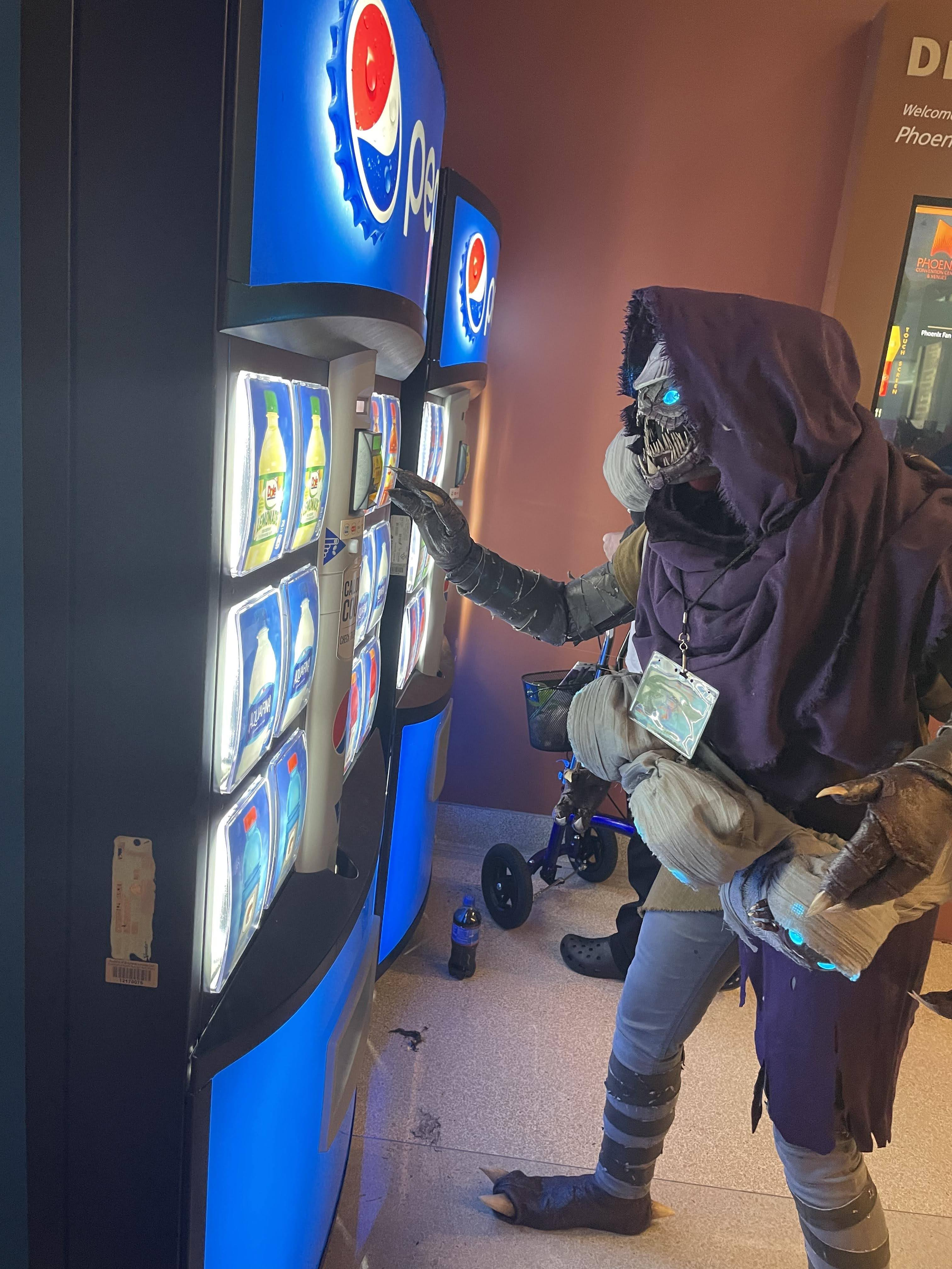 Cosplay of a Buglike alien in a purple hood with 4 glowing blue eyes, 2 claws on each hand, and 2 rows of needle-like teeth. It is trying to buy something from a Pepsi vending machine. It has 4 swaddled babies of the same species in carried in a baby