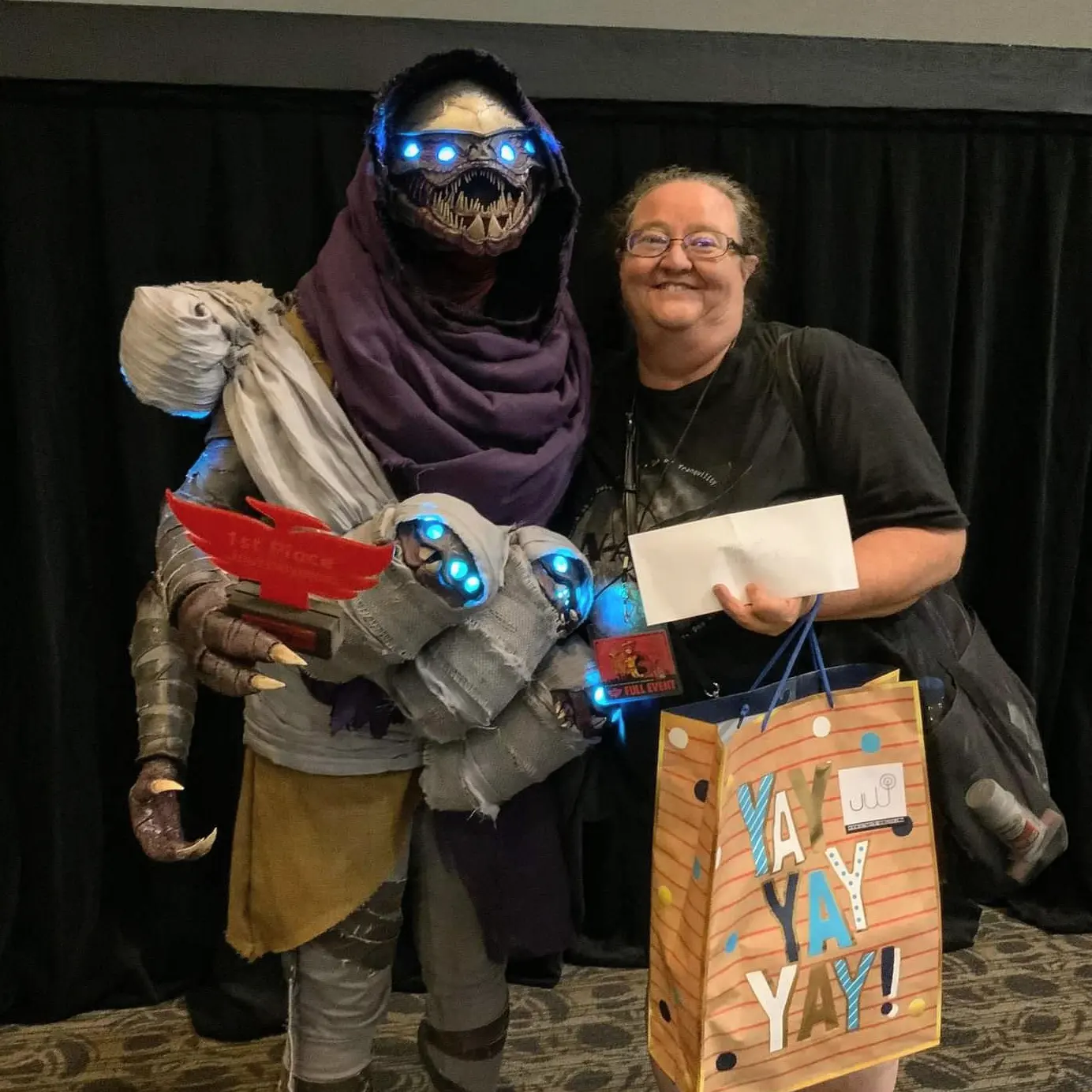 Cosplay of a Buglike alien in a purple hood with 4 glowing blue eyes, 2 claws on each hand, and 2 rows of needle-like teeth. It has 4 swaddled babies of the same species in carried in a baby sling. One hand holds a red Phoenix-shaped cosplay contest 
