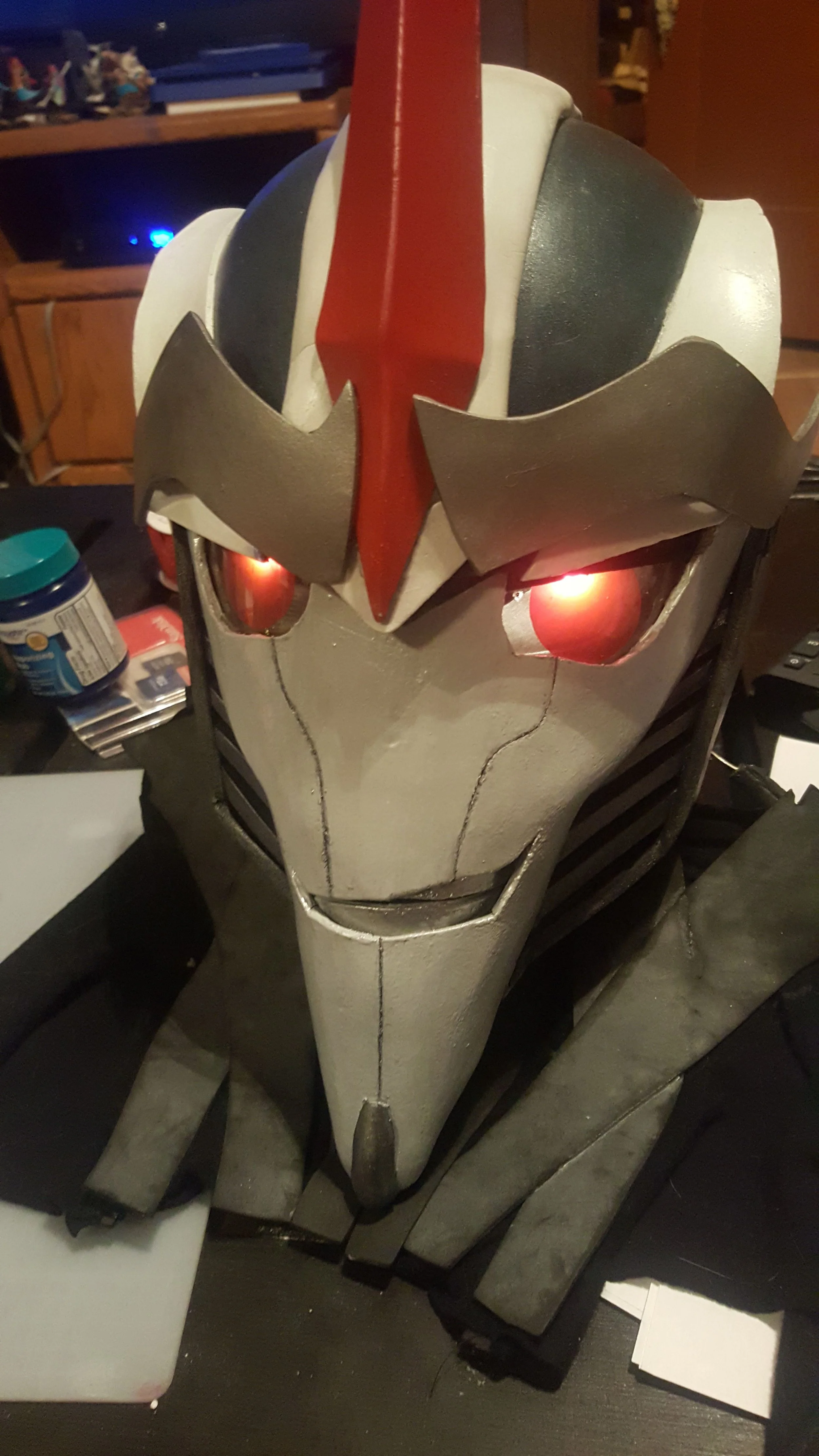 Closeup of Starscream mask with glowing red eyes. The face is pale silver, thin, and pointed, with a smirk. Large silver eyebrows and a red spike adorn the top of the helmet, which is dark blue with white armor strips