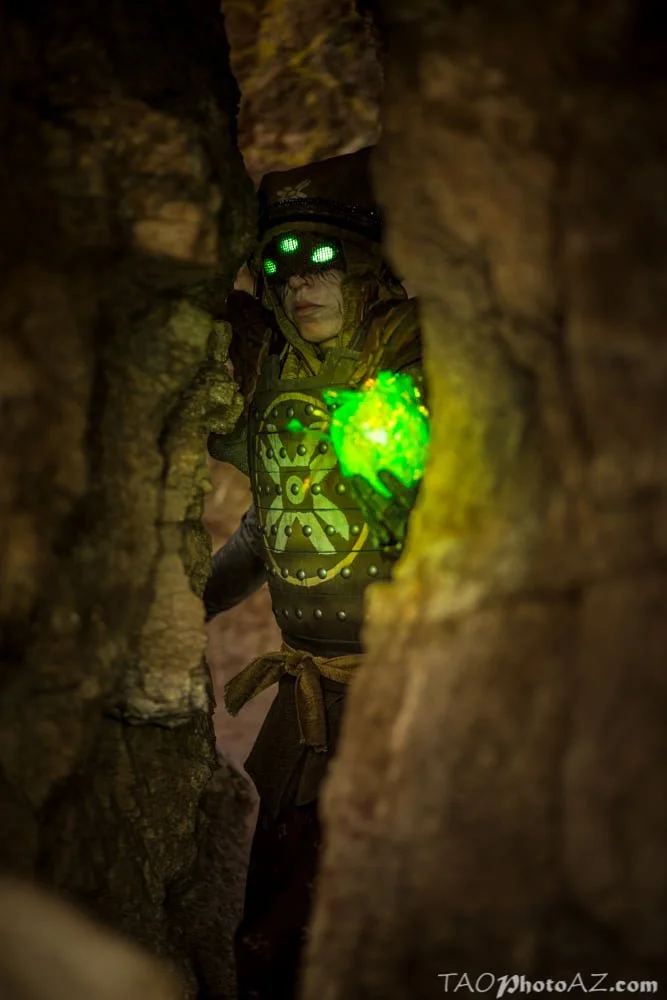 Eris Morn Cosplayer with 3 glowing green eyes holding a glowing green orb as she walks through a cave. The orb's glow shows weathered brown armor and strings of beads and charms. Her face has black tears and a veil with the eyes glowing through