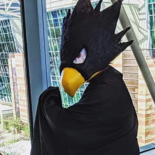 Tokoyami from My Hero Academia Cosplayer in a black cape and black bird head with white eyes and a yellow beak. One fist holds up the cape in a dramatic pose