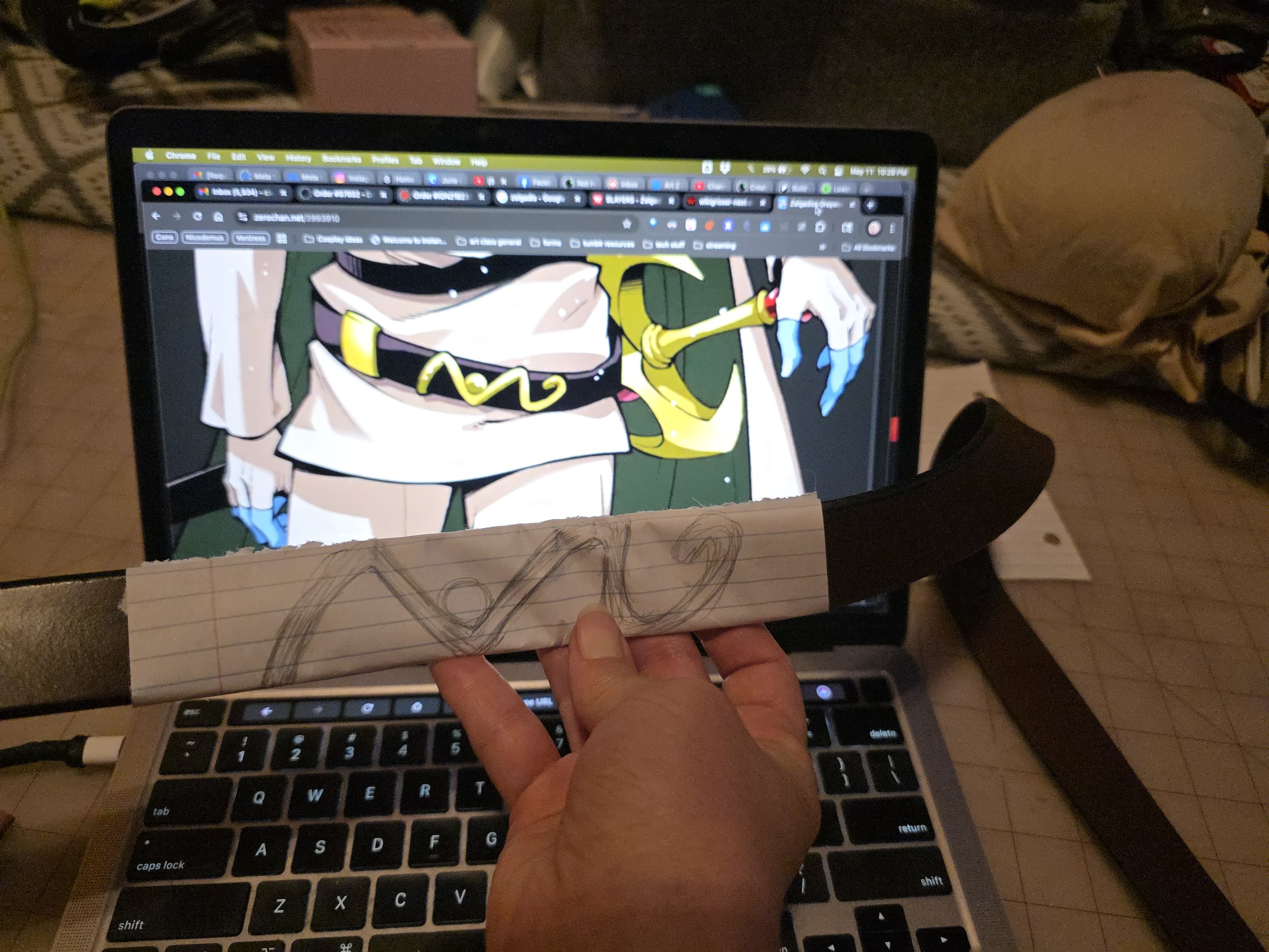 hand holding a sketch of a squiggly shape, next to an image of Zelgadis's sword belts, showing a gold decoration of the same shape.