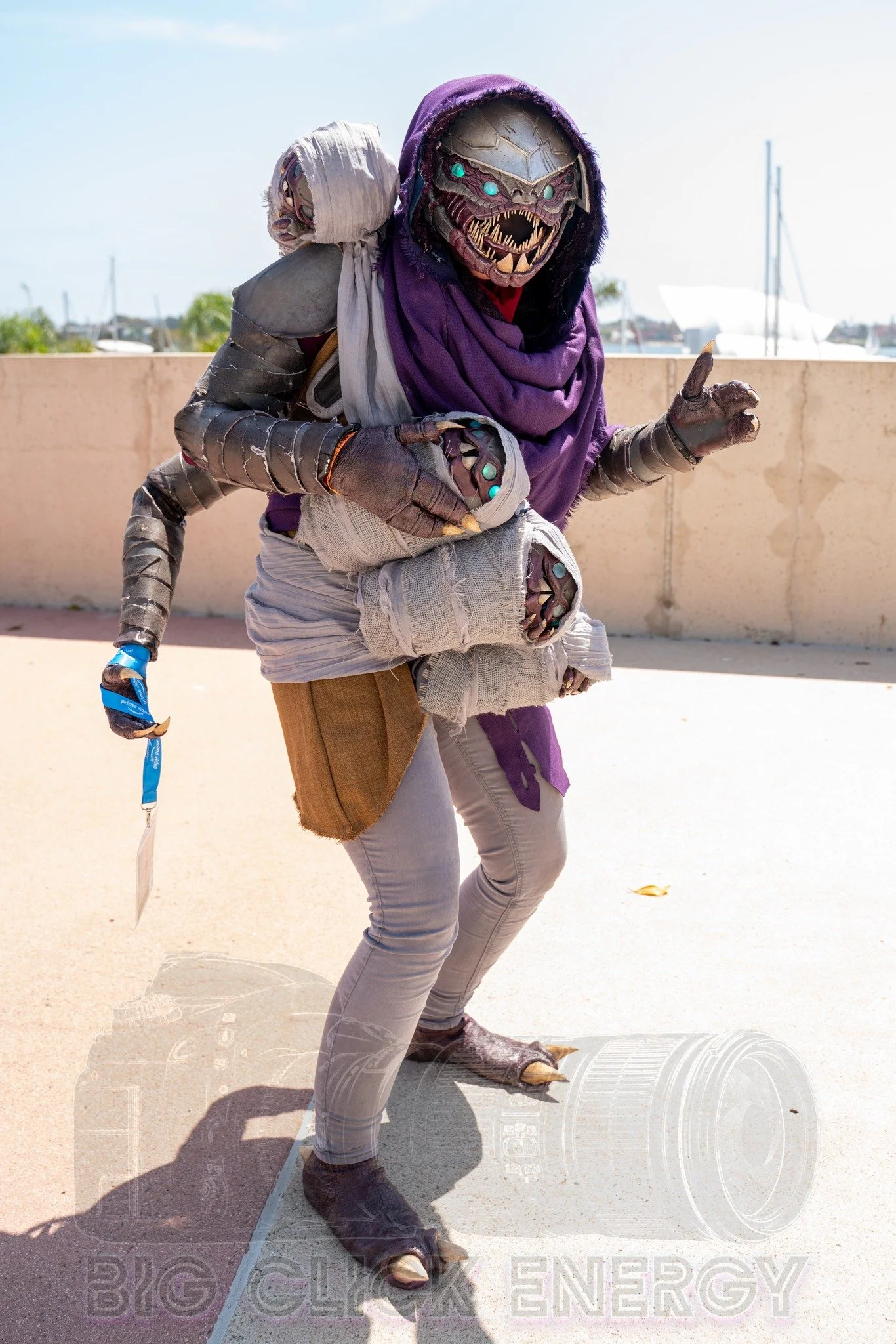 Cosplay of a Buglike alien in a purple hood with 4 glowing blue eyes, 2 claws on each hand, and 2 rows of needle-like teeth. It has 4 swaddled babies of the same species in carried in a baby sling