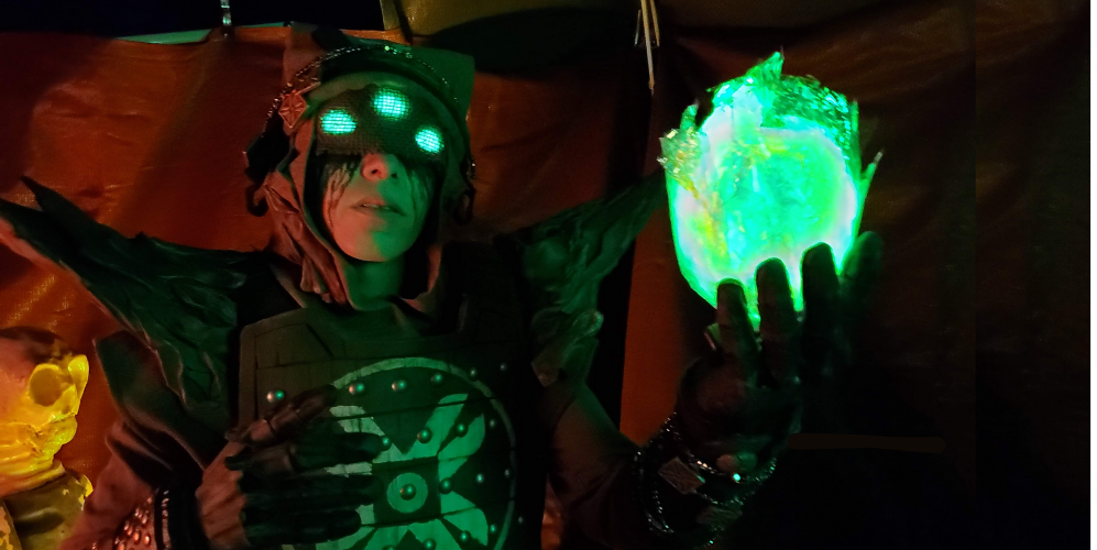 Eris Morn Cosplayer with 3 glowing green eyes holding up a glowing green orb. Black tears flow from under a veil and she wears weathered brown armor. The background is dimly lit and red.