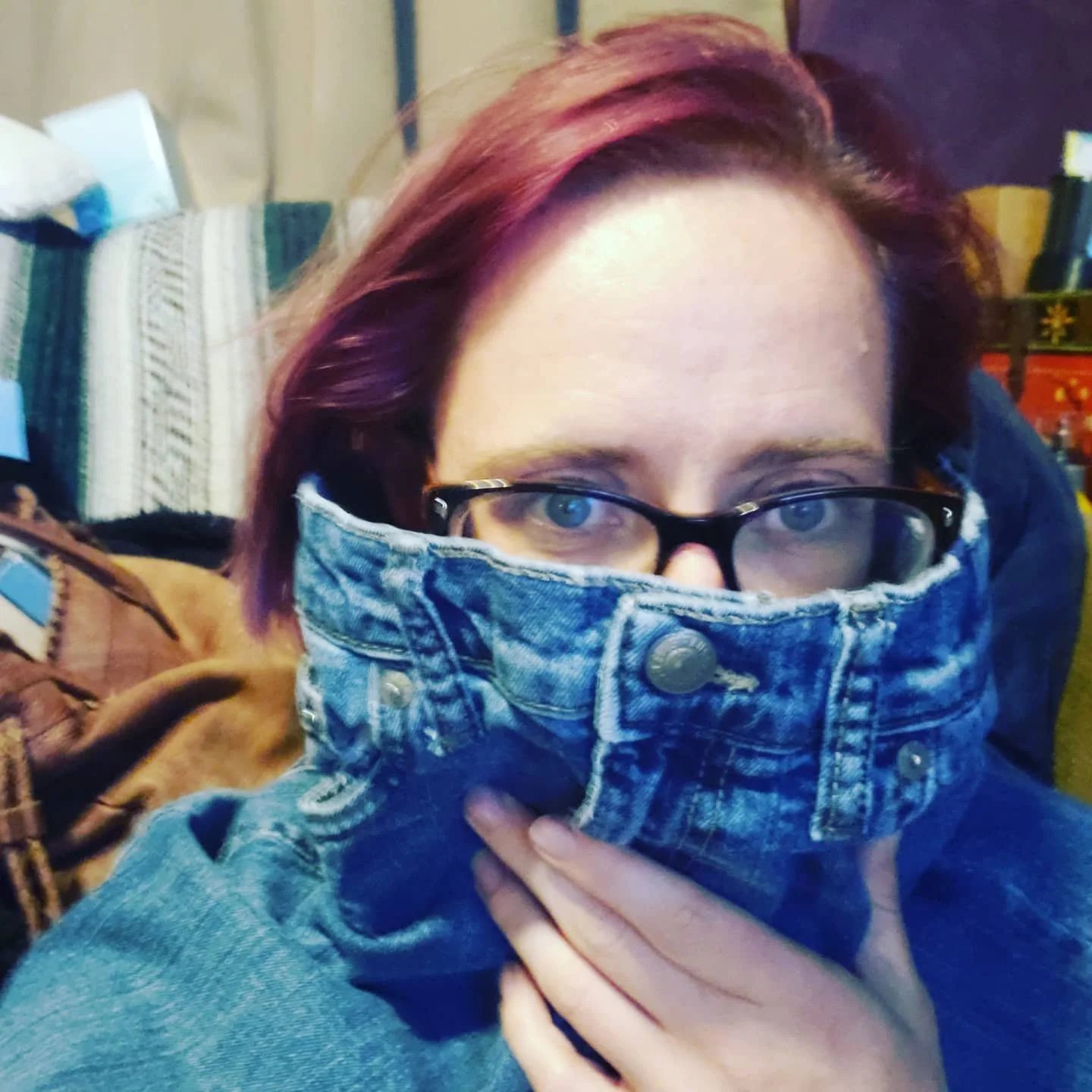 Woman with red hair and black glasses holding the top of some children's jeans up around her face like a mask or high collar