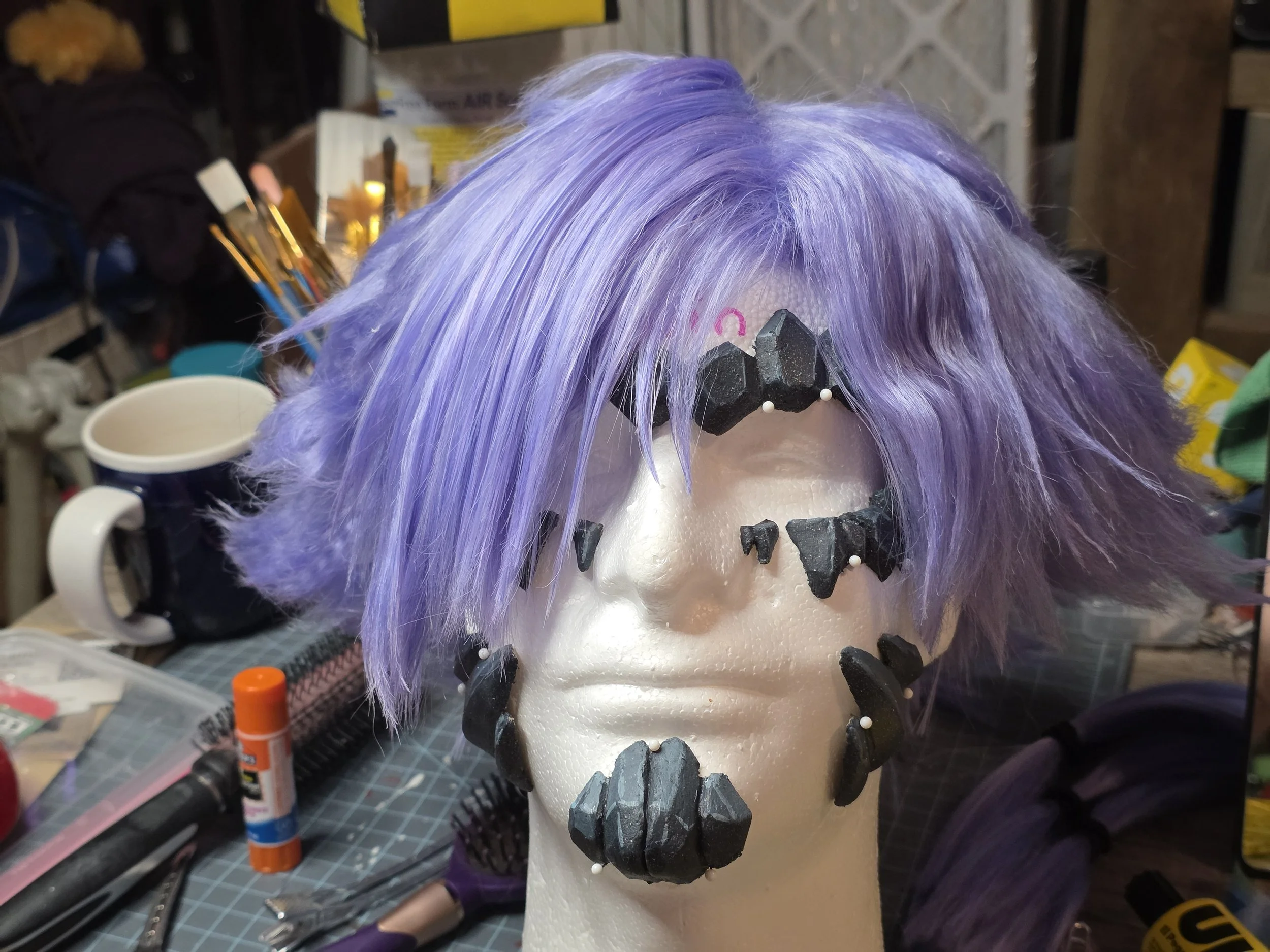 short lavender wig with long bangs combed over one eye. the sides are spiked outwards. Blue-gray, rocklike scales are pinned onto the styrofoam head, along the eyebrows, cheekbones, jawline, and chin