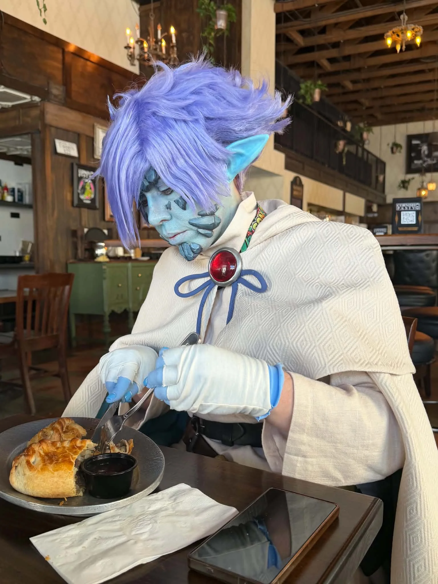 Cosplayer dressed as Zelgadis, an anime character in off-white pants and tunic, off-white cape with dark green lining, blue skin with scale-like rocks in his face, and lavender hair. He sits in a tavern, cutting into a golden brown meat pie with a kn