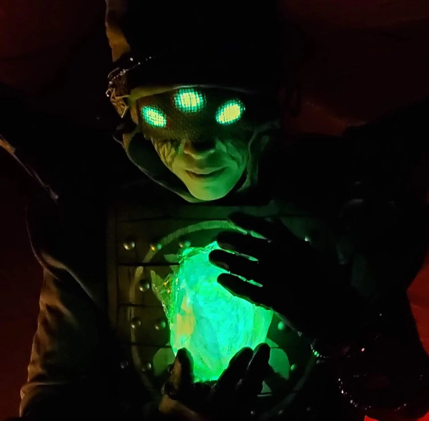 Eris Morn Cosplayer with 3 glowing green eyes clutching a glowing green orb. Black tears flow from under a veil and she wears weathered brown armor. The background is dark
