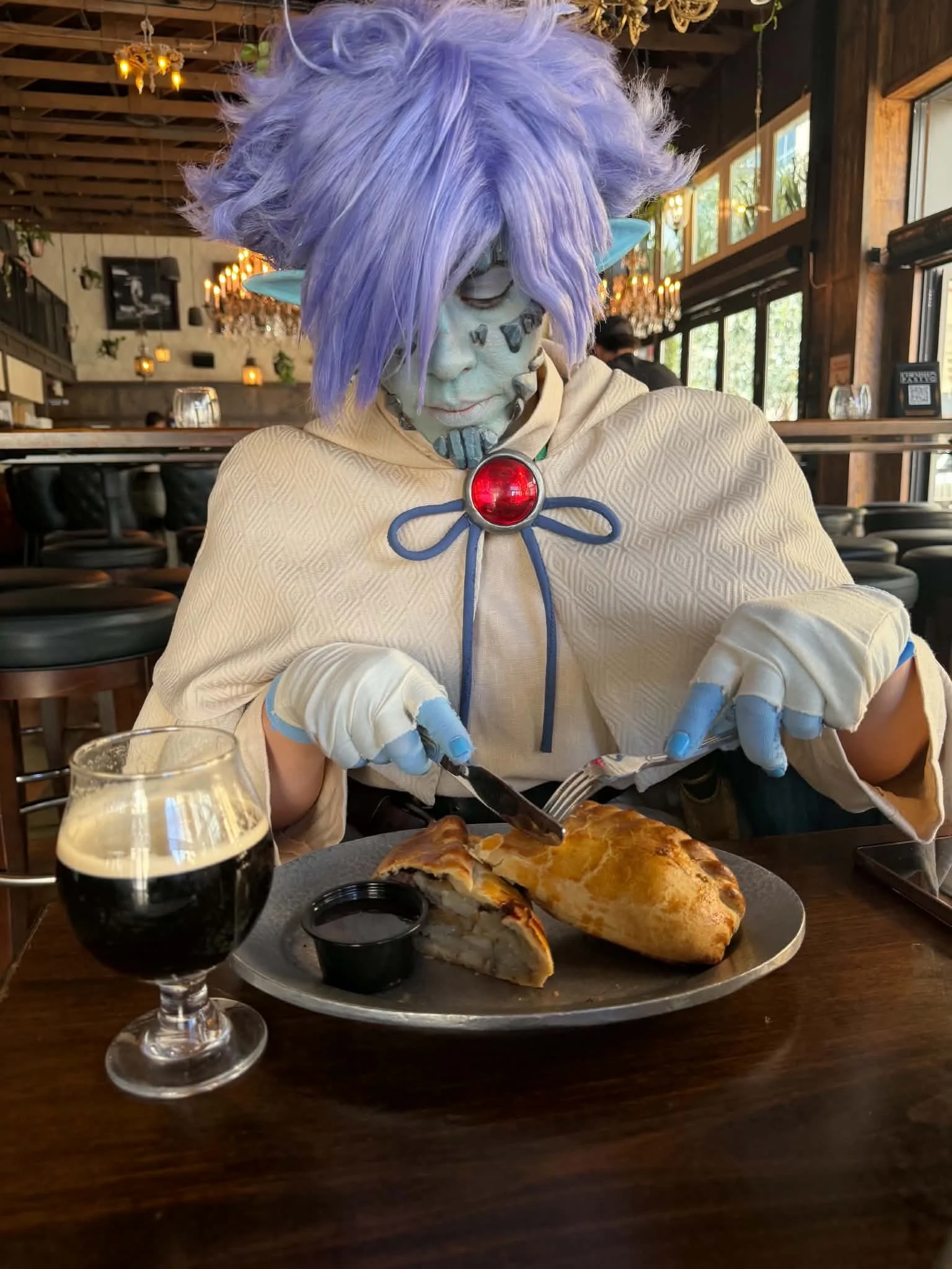 Cosplayer dressed as Zelgadis, an anime character in off-white pants and tunic, off-white cape with dark green lining, blue skin with scale-like rocks in his face, and lavender hair. He sits in a tavern, cutting into a golden brown meat pie with a kn