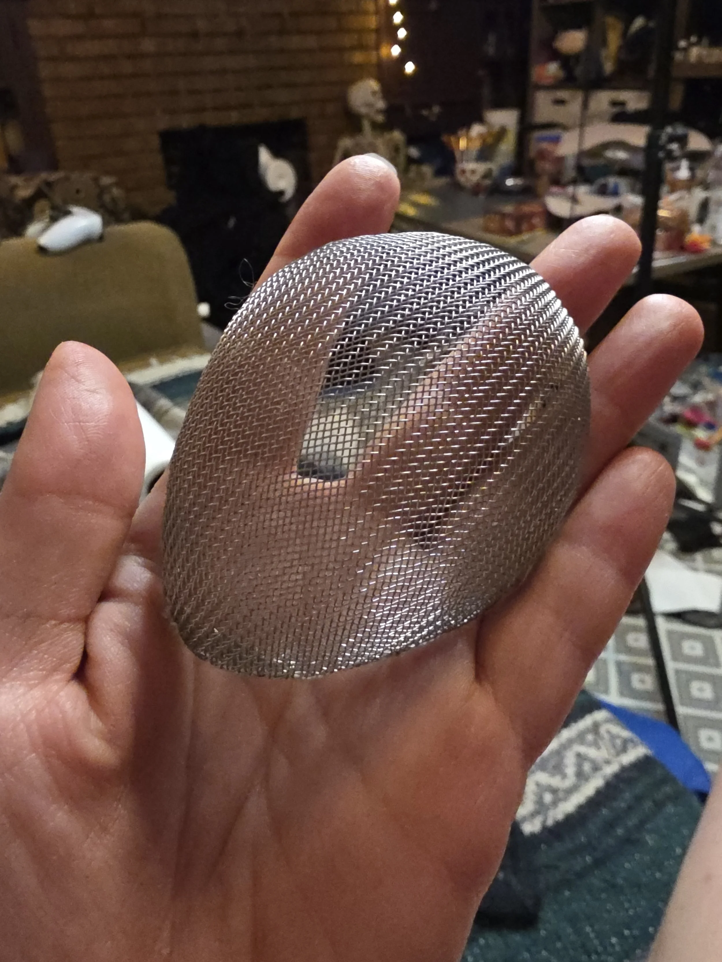 Metal screen kitchen strainer, but the frame and handle have been removed. A hand is holding just the strainer part, bent into a half egg shape. Fingers are visible behind the mesh.
