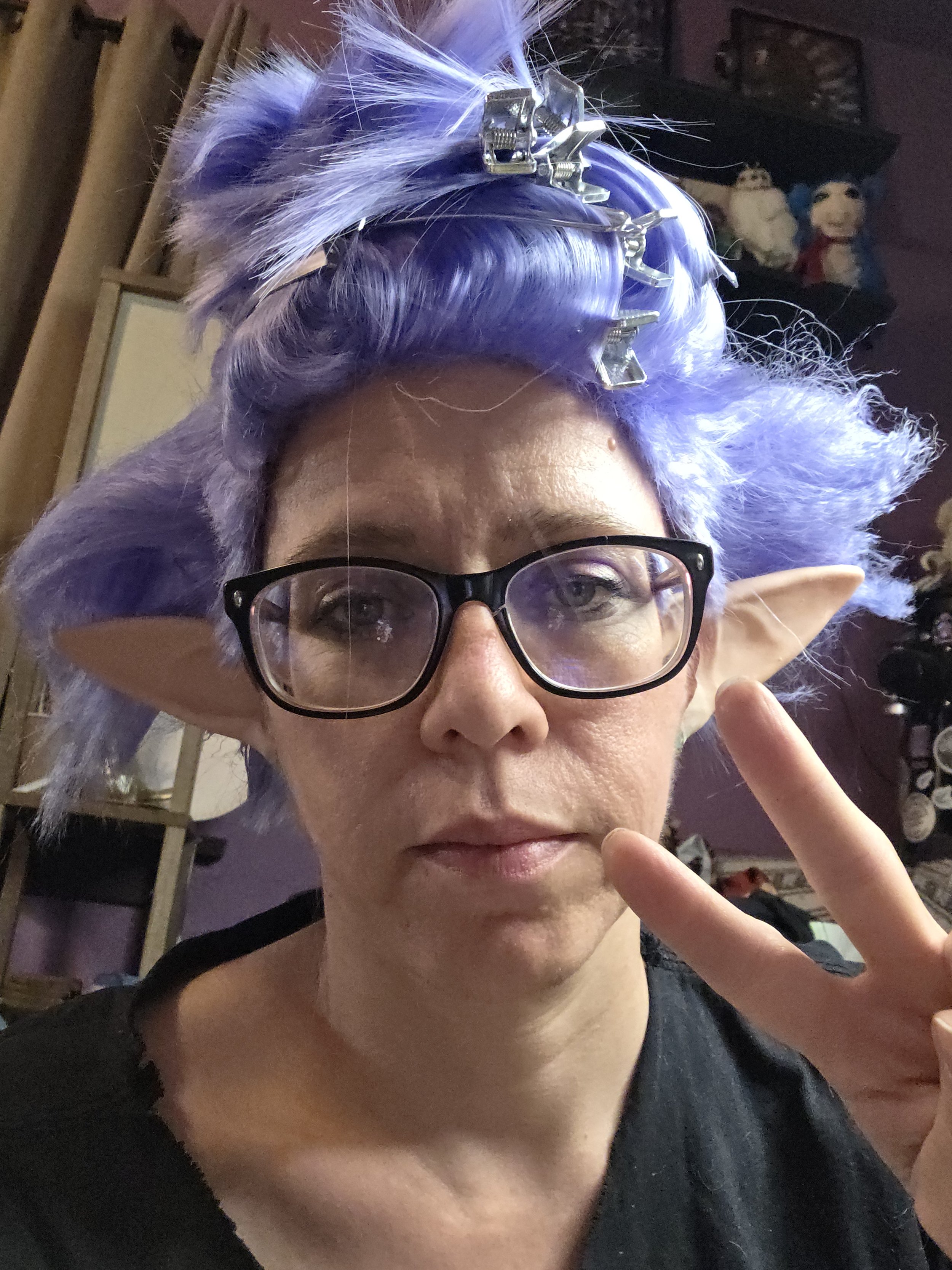 cosplayer with a tired look showing a peace sign, with a lavender wig on her head. half the wig is pinned up and half is teased and frizzy