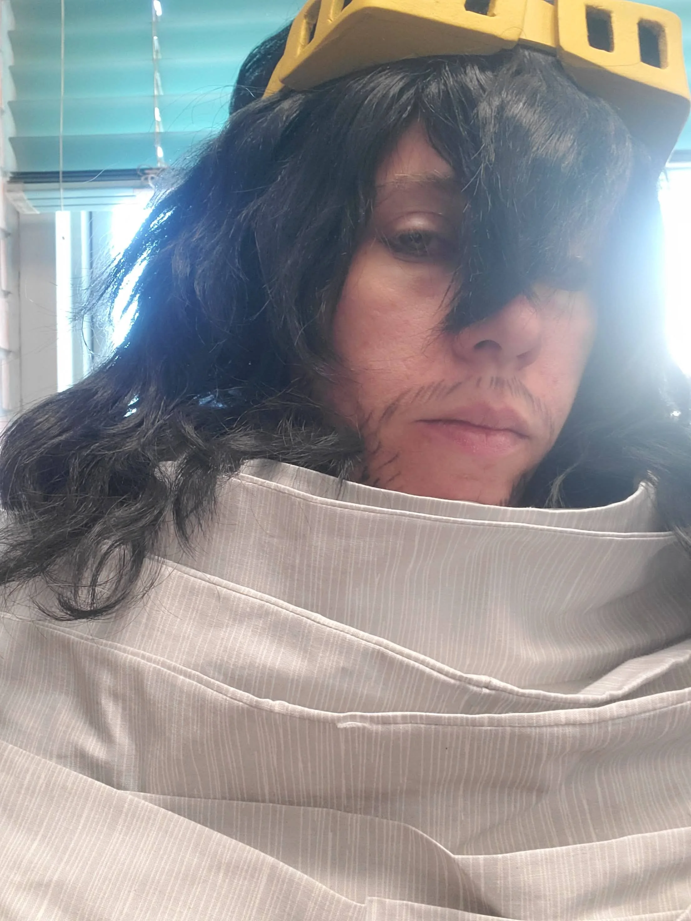 Cosplay of Aizawa from My Hero Academia in a messy midlength black wig, yellow goggles, and a long white scarf wrapped many times around the shoulders. She has a tired expression and has a scraggly beard and moustache drawn on with eyeliner