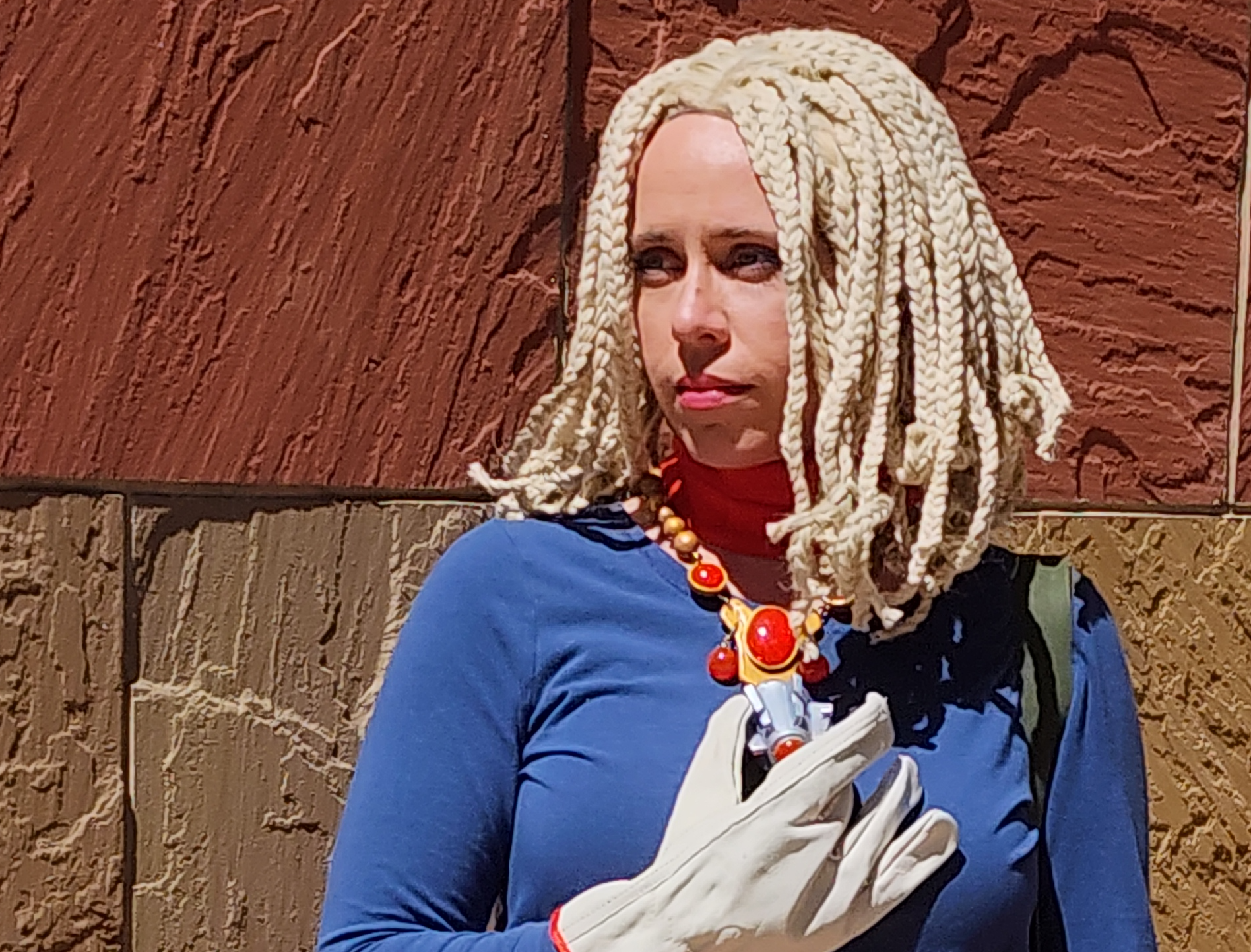 Cosplayer in a light blond braided wig, blue long sleeved shirt, and work gloves. She has an elaborate gold necklace with red resin gems