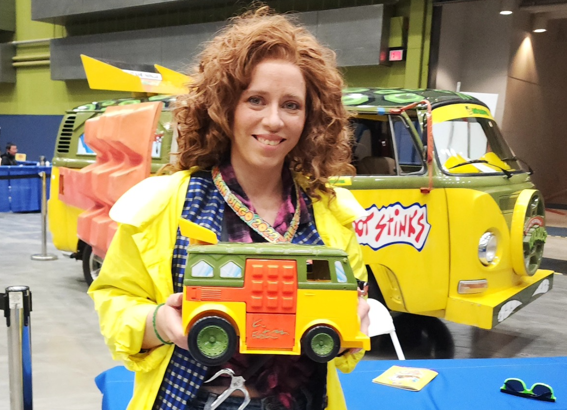 Cosplayer dressed in curly reddish wig, yellow raincoat, patterned vest, with a sai in her belt. She smiles holding a toy Turtle Van while standing in front of a life size Turtle Van