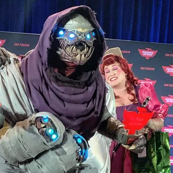 Cosplay of a Buglike alien in a purple hood with 4 glowing blue eyes, 2 claws on each hand, and 2 rows of needle-like teeth. It has 4 swaddled babies of the same species in carried in a baby sling. One hand holds a red Phoenix-shaped cosplay contest 