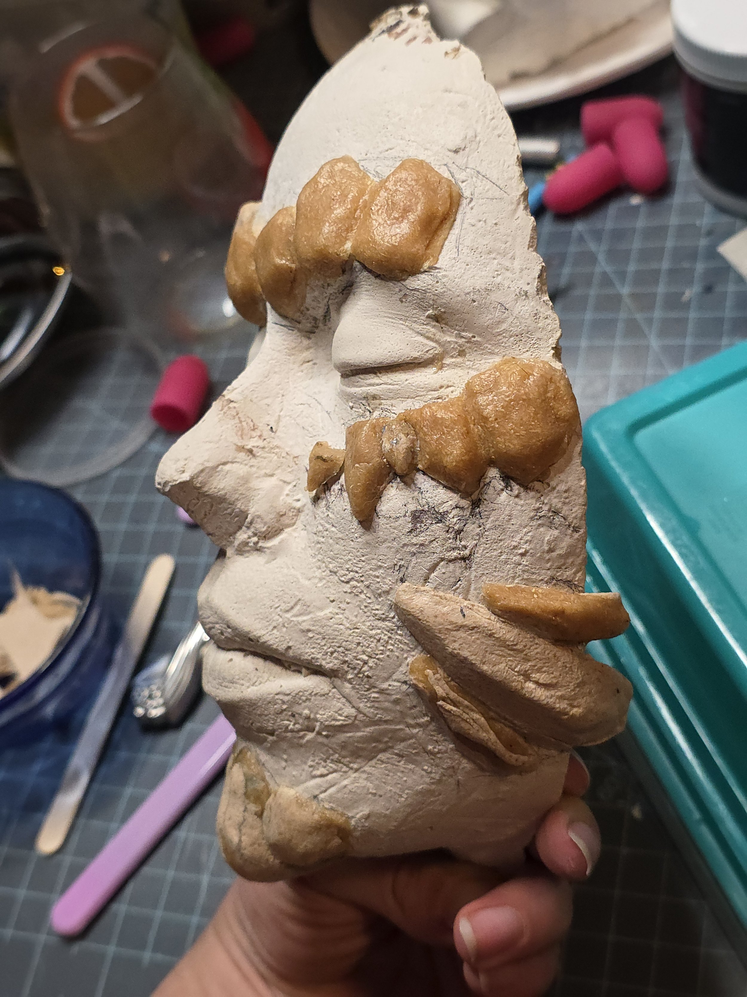 side view plaster cast of face with pieces of tan colored Worbla stuck on to form rock-like scales. The eyebrows, cheekbones, and jawline have mostly flat scales with some built-up thickness, and the chin has a goatee like shape made up worbla rocks