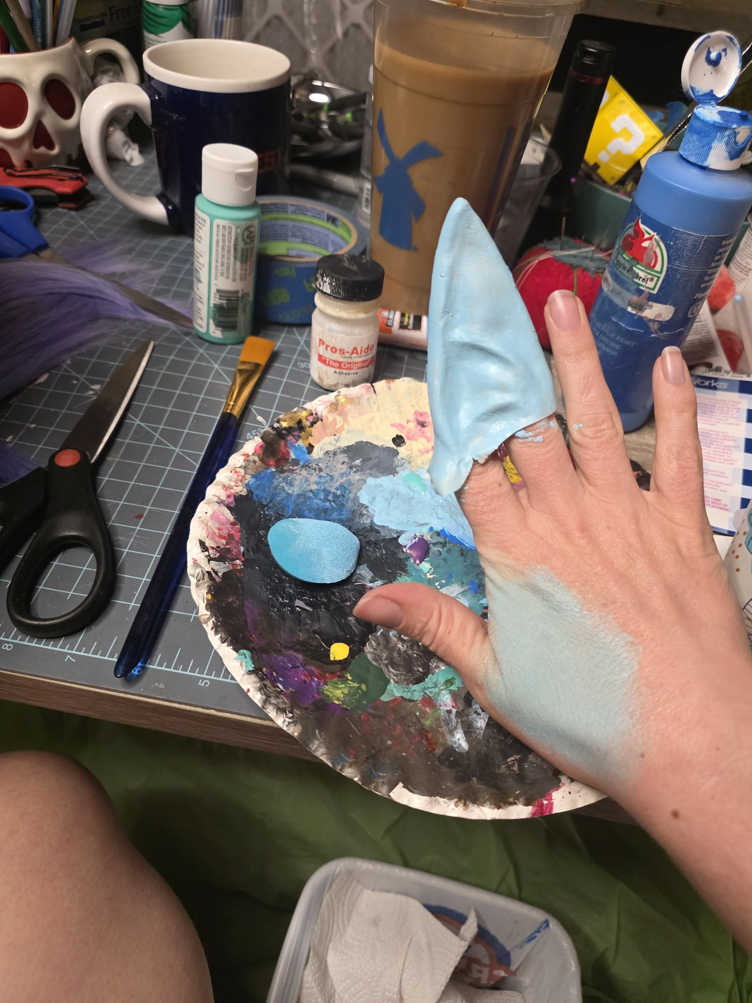 latex pointy ear held on 2 fingers, painted pale blue. paint is mixed on a paper plate, with a bottle of Pros-Aide, because that is how paint sticks to latex. Pale blue makeup is swatched on the hand holding the ear, and is close color match for the 