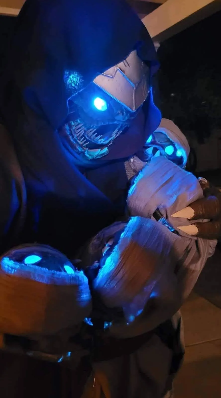 Cosplay of a Buglike alien in a purple hood with 4 glowing blue eyes, 2 claws on each hand, and 2 rows of needle-like teeth. It cradles 4 swaddled babies of the same species in its lap