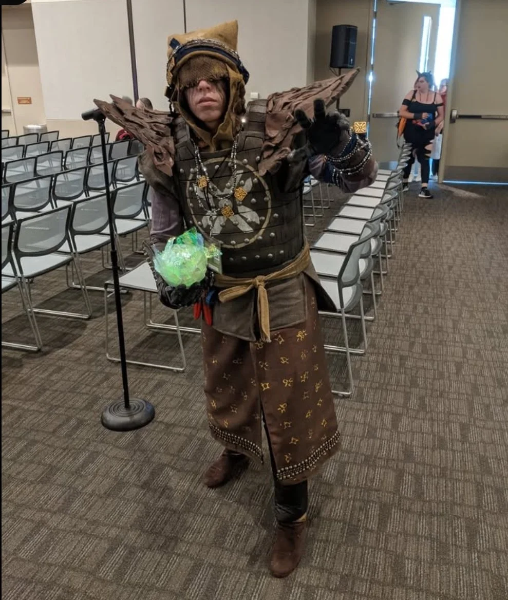 Eris Morn Cosplayer with 3 glowing green eyes holding a glowing green orb in a room full of chairs at a comic con. She wears weathered brown armor and strings of beads and charms. Her face has black tears and a veil with the eyes glowing through