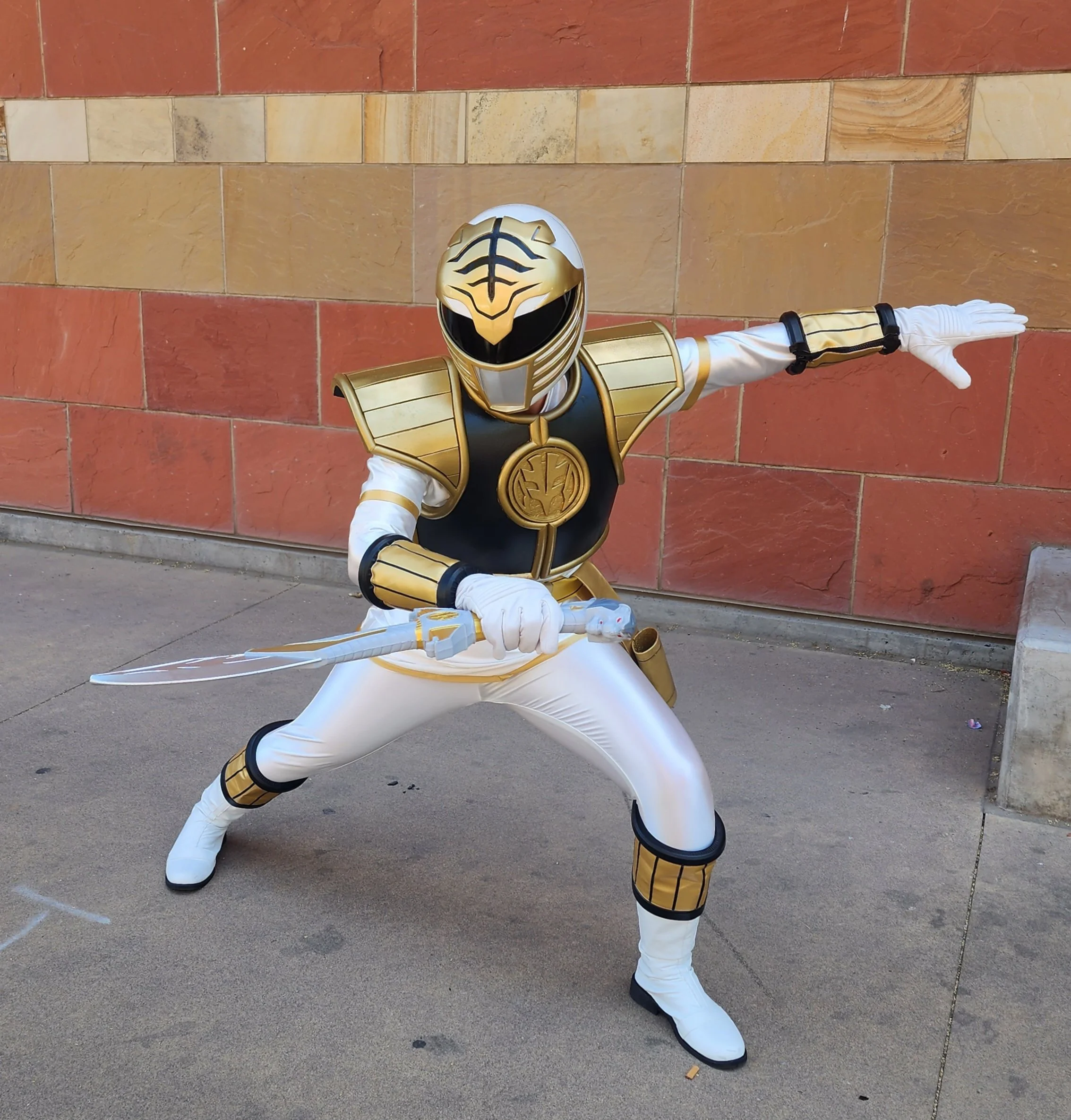 Cosplay of the White Ranger standing in a martial arts stance holding a curved sword with a tiger handle. The suit is white and shiny, with gold and black armor and cuffs. The gold and white helmet resembles a tiger