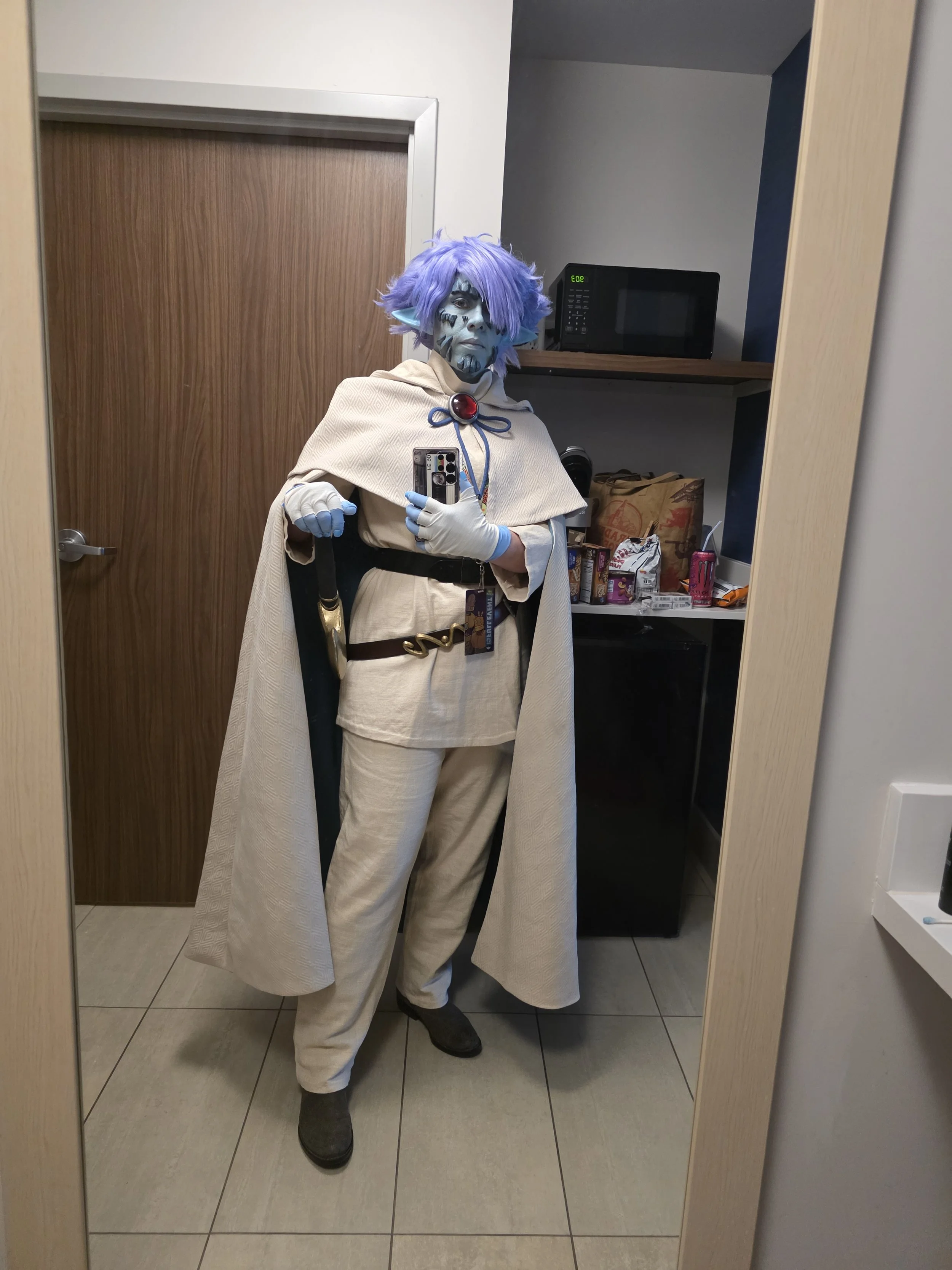 Cosplayer dressed as Zelgadis, an anime character in off-white pants and tunic, off-white cape with dark green lining, blue skin with scale-like rocks in his face, and lavender hair. His hand rests on the hilt of a sword hung from one of two belts on