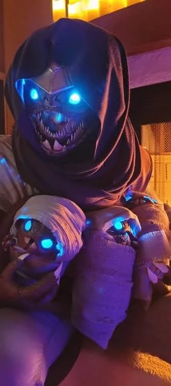 Cosplay of a Buglike alien with 4 glowing blue eyes, 2 claws on each hand, and 2 rows of needle-like teeth. It has 4 swaddled babies of the same species, wears a purple hood