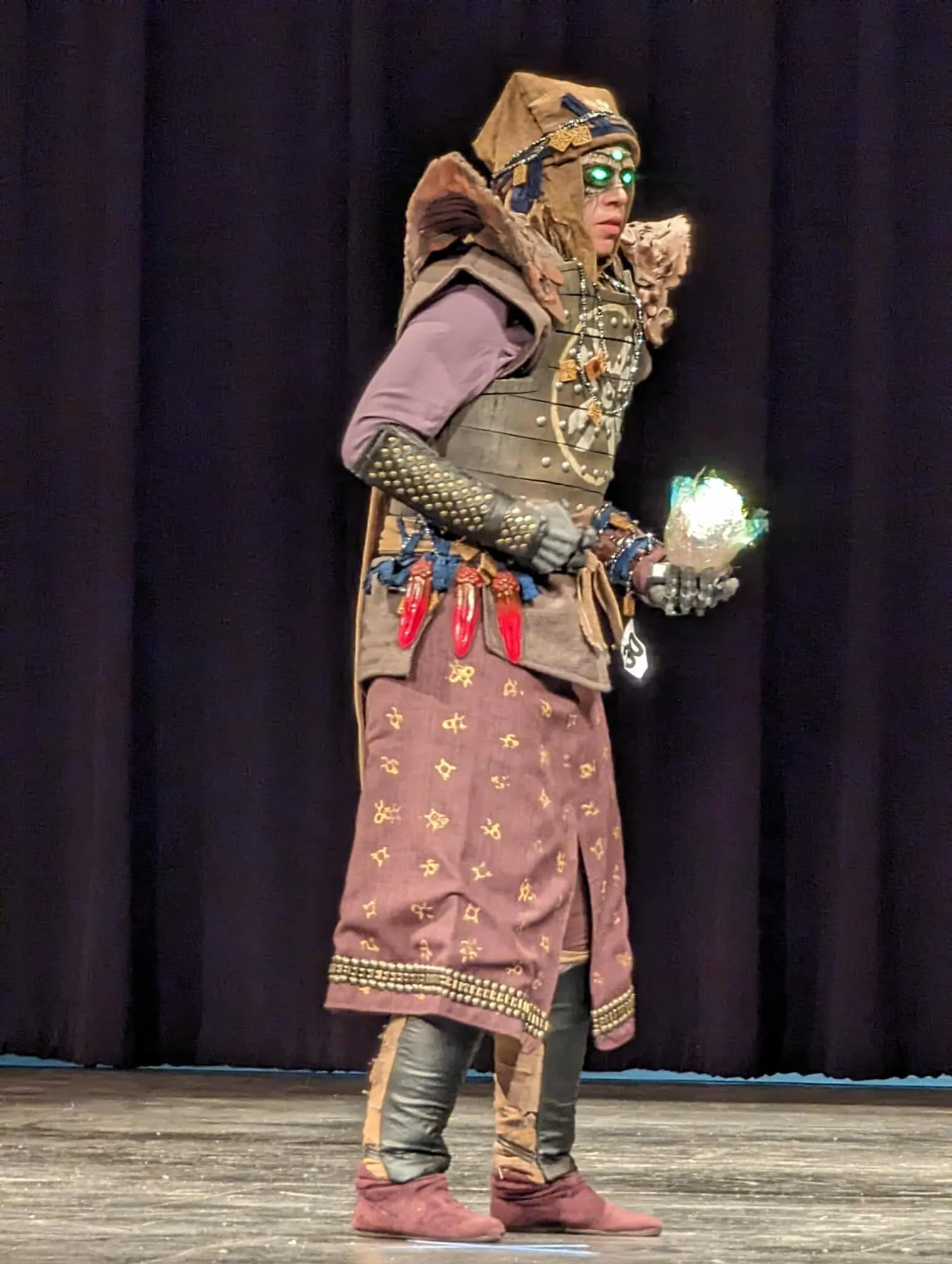 Side view of an Eris Morn Cosplayer with 3 glowing green eyes holding a glowing green orb. She wears weathered brown armor and strings of beads and charms. Her face has monstrous features and scars around the 3 eyes