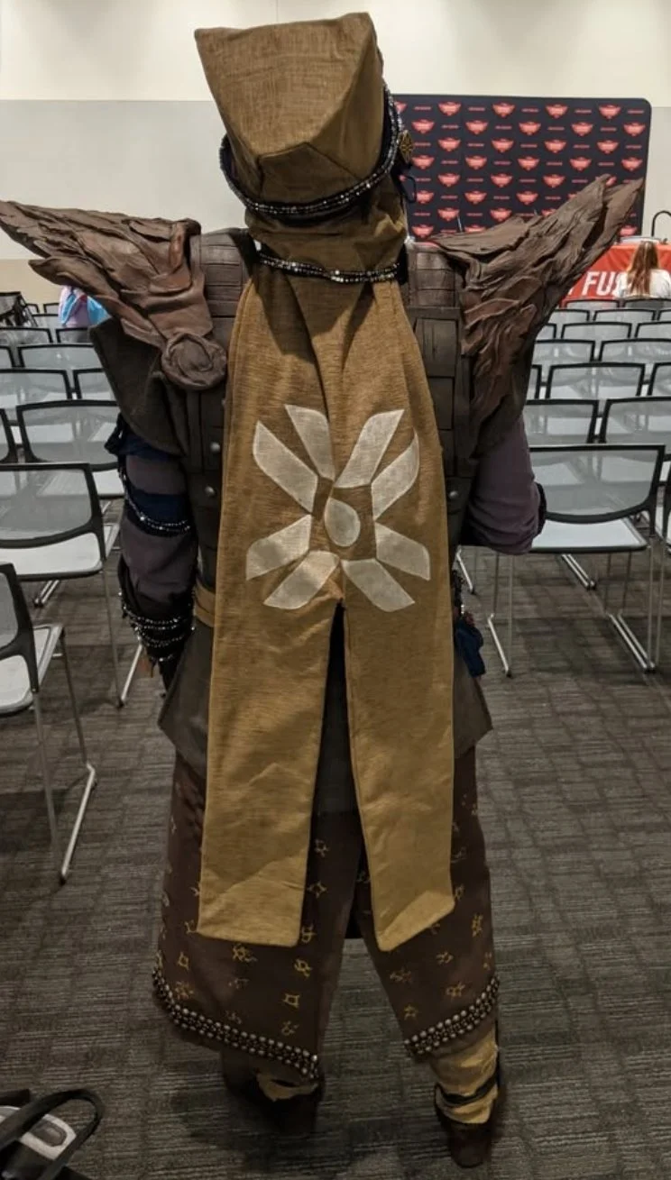 Back view of an Eris Morn Cosplayer in a room full of chairs at a comic con. She wears a narrow tan cape with a flower-like symbol in the middle, weathered brown armor and strings of beads and charms. 