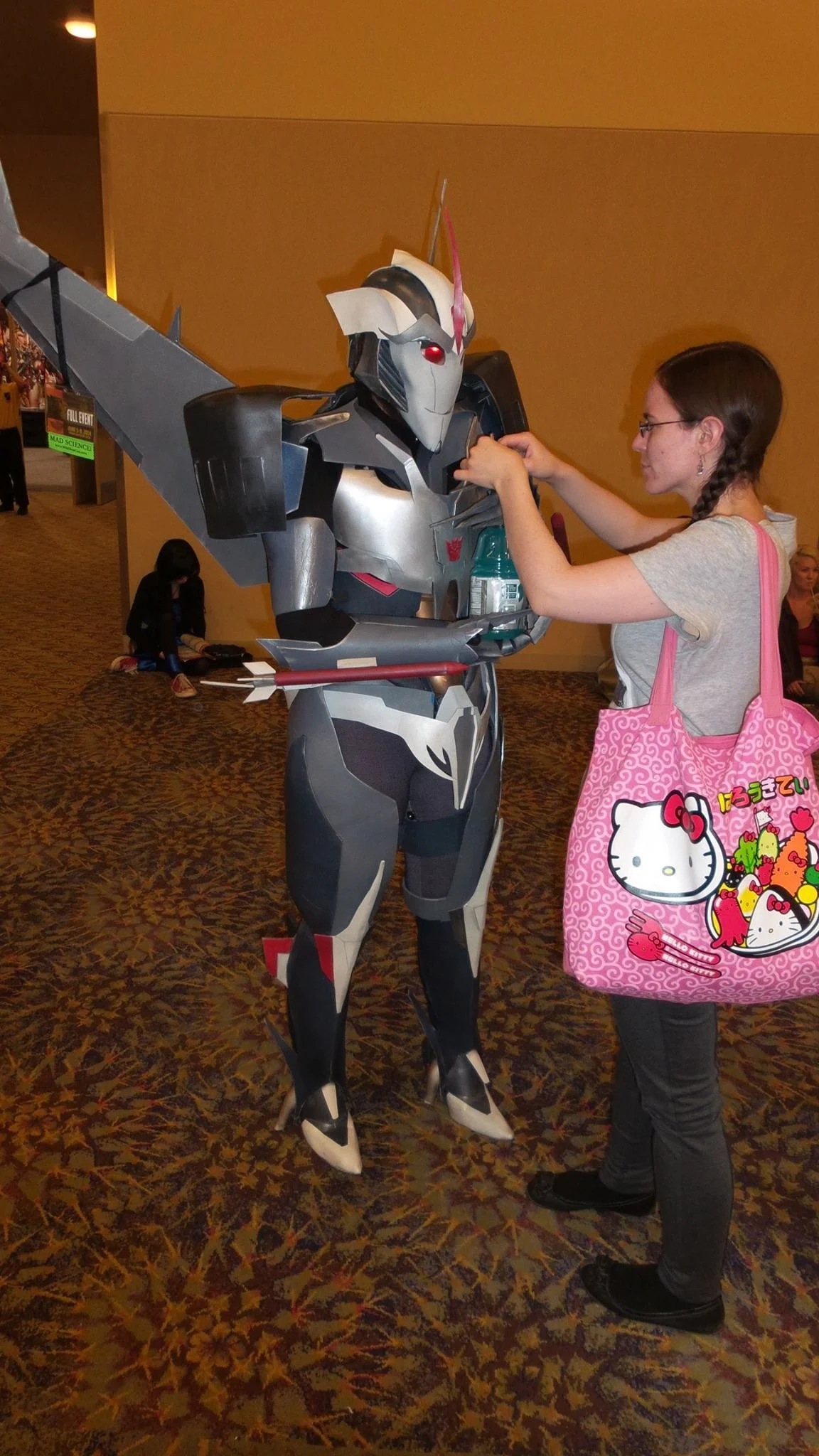 Young woman with a large pink Hello Kitty bag holds a straw so a Cosplayer in a complicated Starscream suit can drink blue gatorade. The cosplay is gray and silver armor with a mask and large upward pointing wings