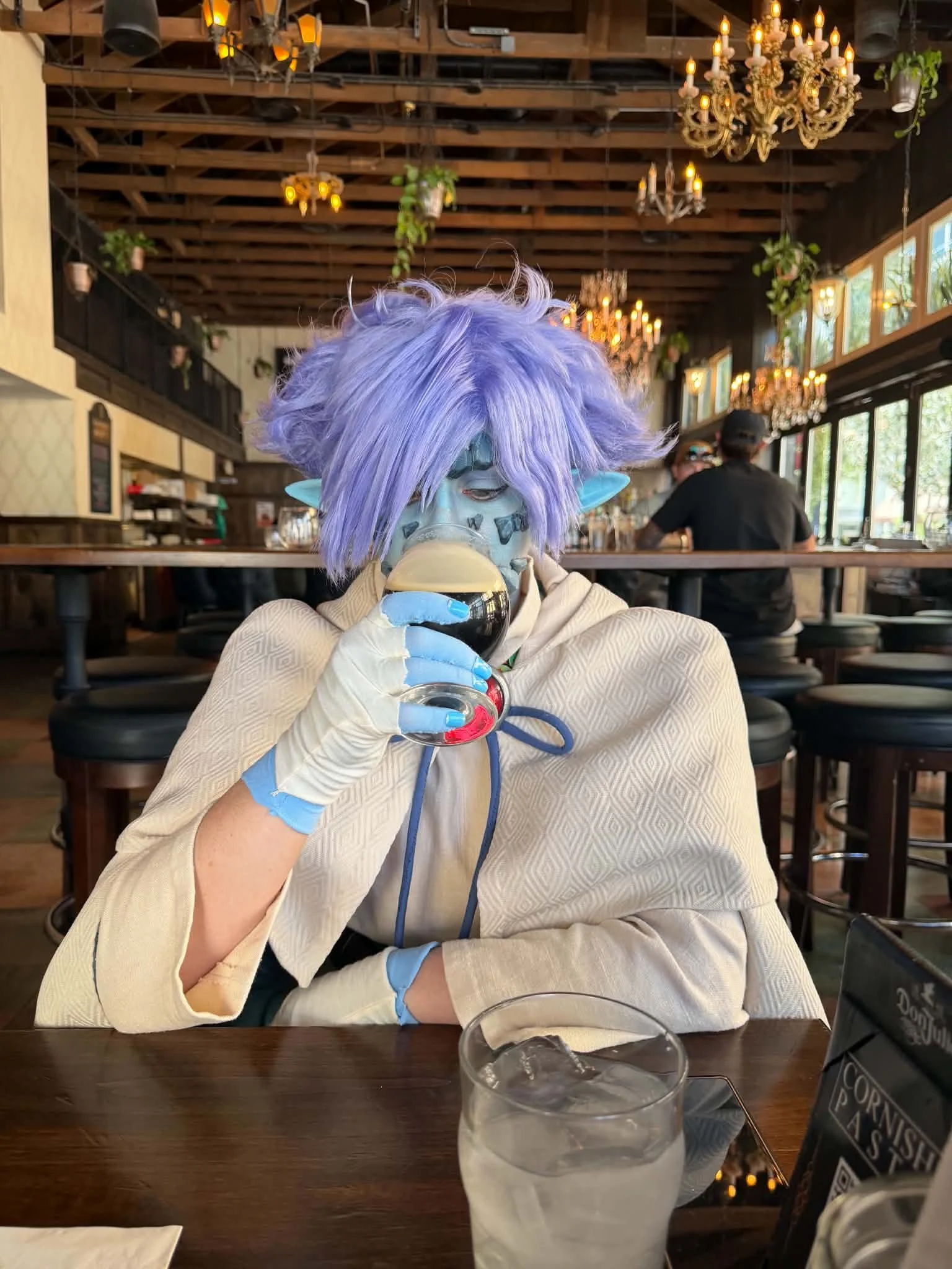 Cosplayer dressed as Zelgadis, an anime character in off-white pants and tunic, off-white cape with dark green lining, blue skin with scale-like rocks in his face, and lavender hair. He sits in a tavern, eyes closed, drinking from a glass of brown be
