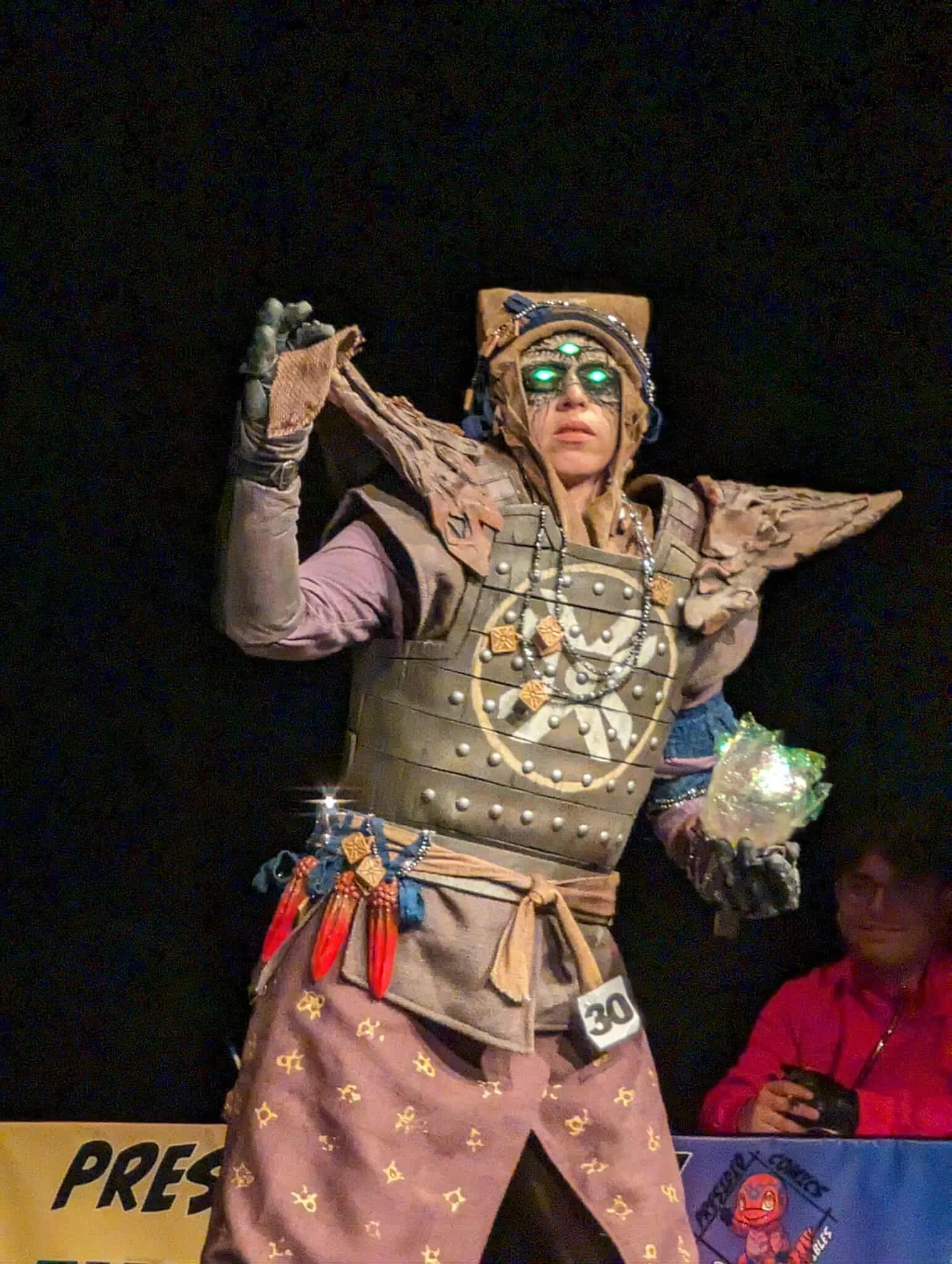 Eris Morn Cosplayer with 3 glowing green eyes holding a glowing green orb and reaching out with the other hand. She wears weathered brown armor and strings of beads and charms. Her face has monstrous features and scars around the 3 eyes