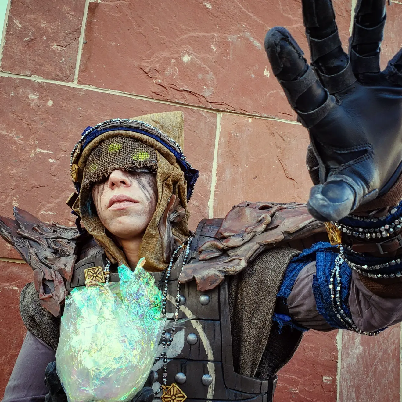 Eris Morn Cosplayer with 3 glowing green eyes holding a glowing green orb and reaching out with the other hand. She wears weathered brown armor and strings of beads and charms. Her face has black tears and a veil with the eyes glowing through