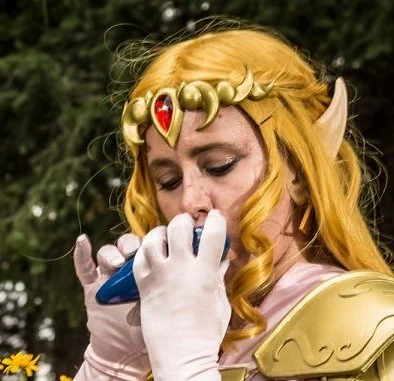 Cosplayer dressed as Zelda in a golden blond wig, gold crown with a red gem, gold armor, and a pink dress and gloves. She looks down while playing a blue ocarina