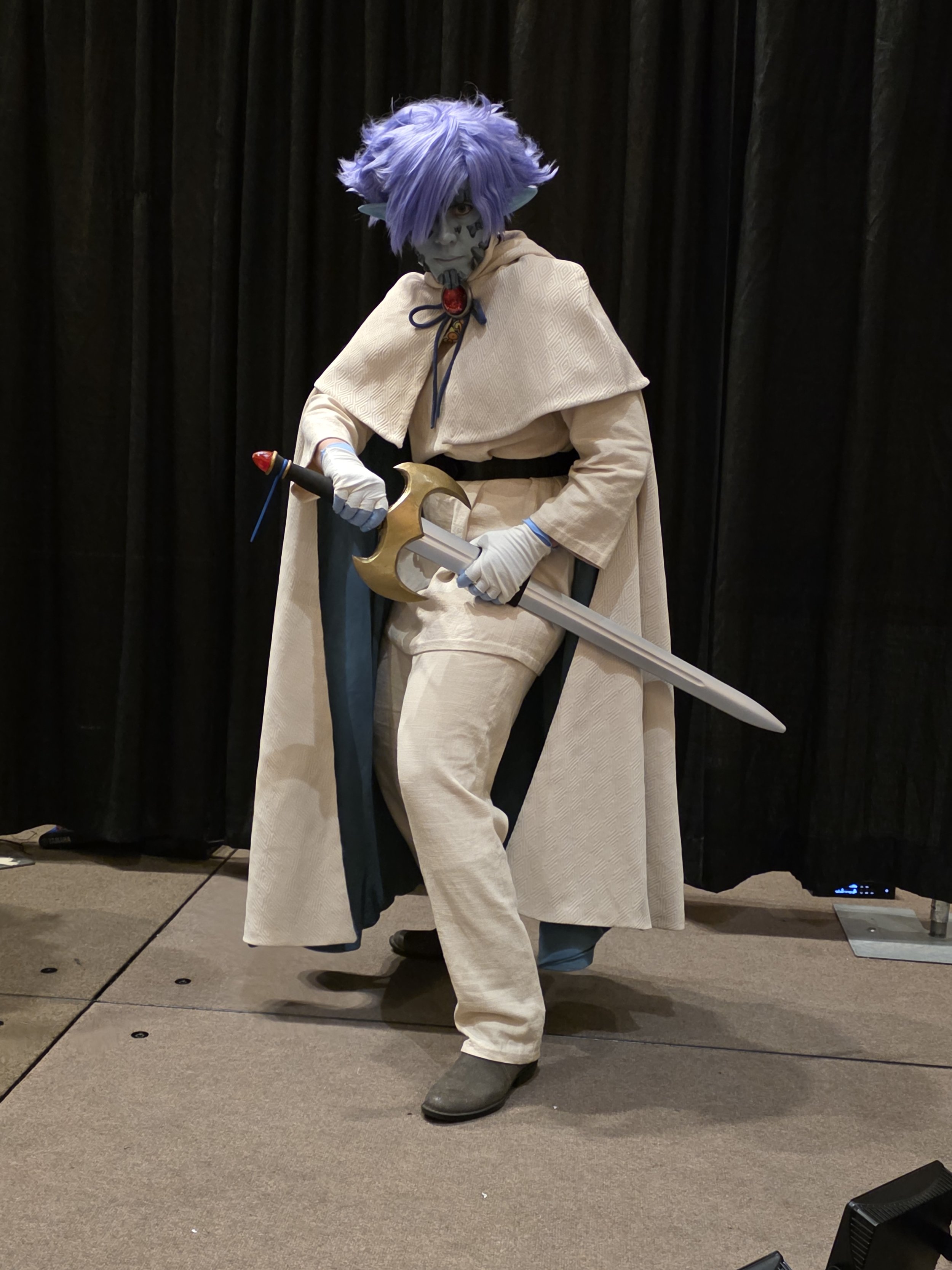 Cosplayer dressed as Zelgadis, an anime character in off-white pants and tunic, off-white cape with dark green lining, blue skin with scale-like rocks in his face, and lavender hair. He is about to draw a longsword with a large golden hilt