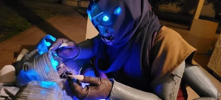 Cosplay of a Buglike alien in a purple hood and brown tunic, with 4 glowing blue eyes, 2 claws on each hand, and 2 rows of needle-like teeth. It holds the hand of a swaddled baby of the same species