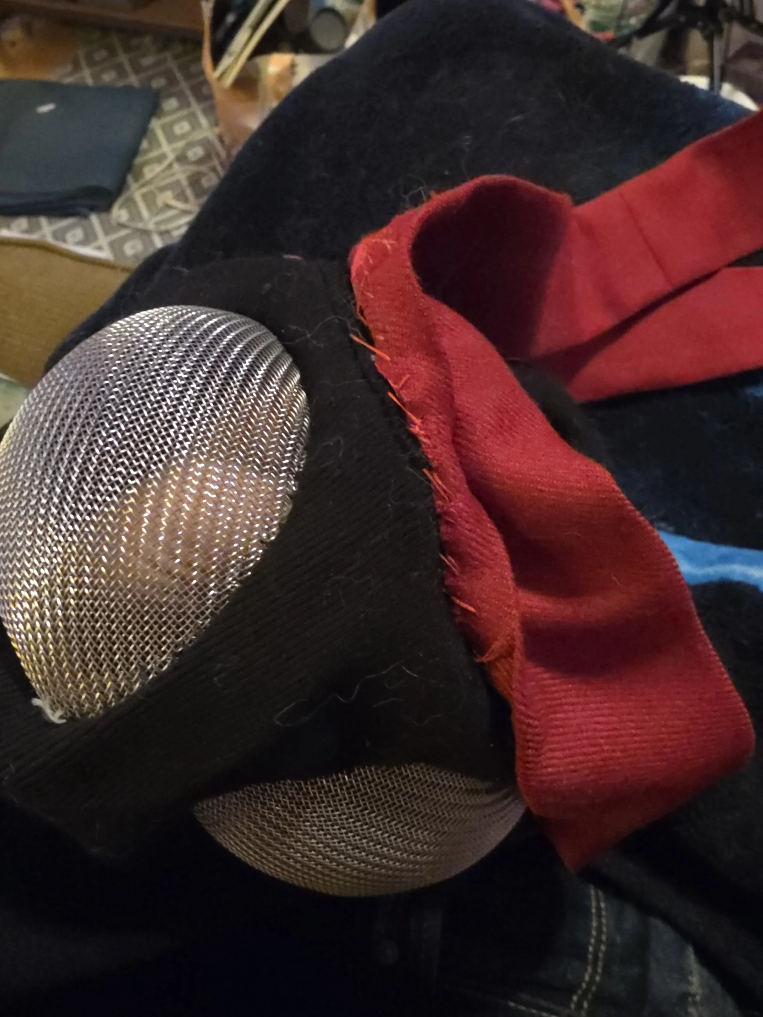 Horehead area of a black fabric ninja mask with metal screen eye covers. A red headband if flipped up to show where it is being stitched onto the mask above the eyes