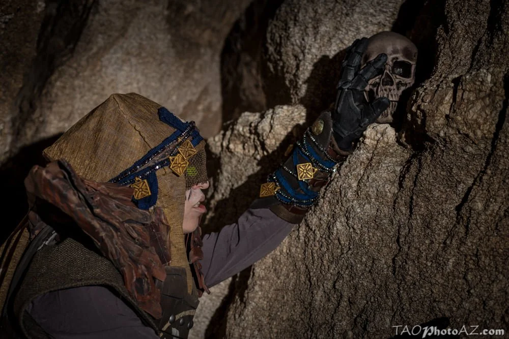Eris Morn Cosplayer with 3 glowing green eyes reaching up for a skull on a rock shelf in a cave. She wears weathered brown armor and strings of beads and charms. Her face has black tears and a veil with the eyes glowing through