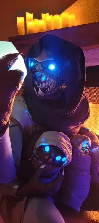 Cosplay of a Buglike alien with 4 glowing blue eyes, 2 claws on each hand, and 2 rows of needle-like teeth. It has swaddled babies of the same species, wears a purple hood, and holds up a yellow glowing dodecahedron
