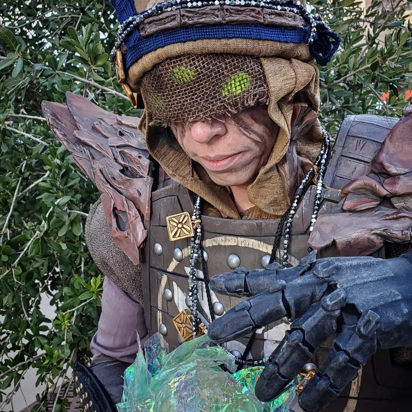 Eris Morn Cosplayer with 3 glowing green eyes holding a glowing green orb. She wears gray armored gloves, weathered brown armor and strings of beads and charms. Her face has black tears and a veil with the eyes glowing through